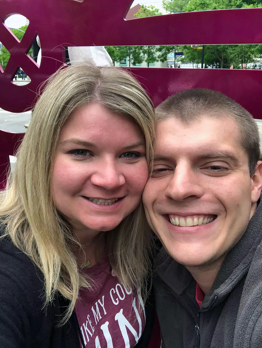 Katie and Zack smiling in a selfie taken in New York City, with urban scenery and iconic landmarks softly visible in the background.