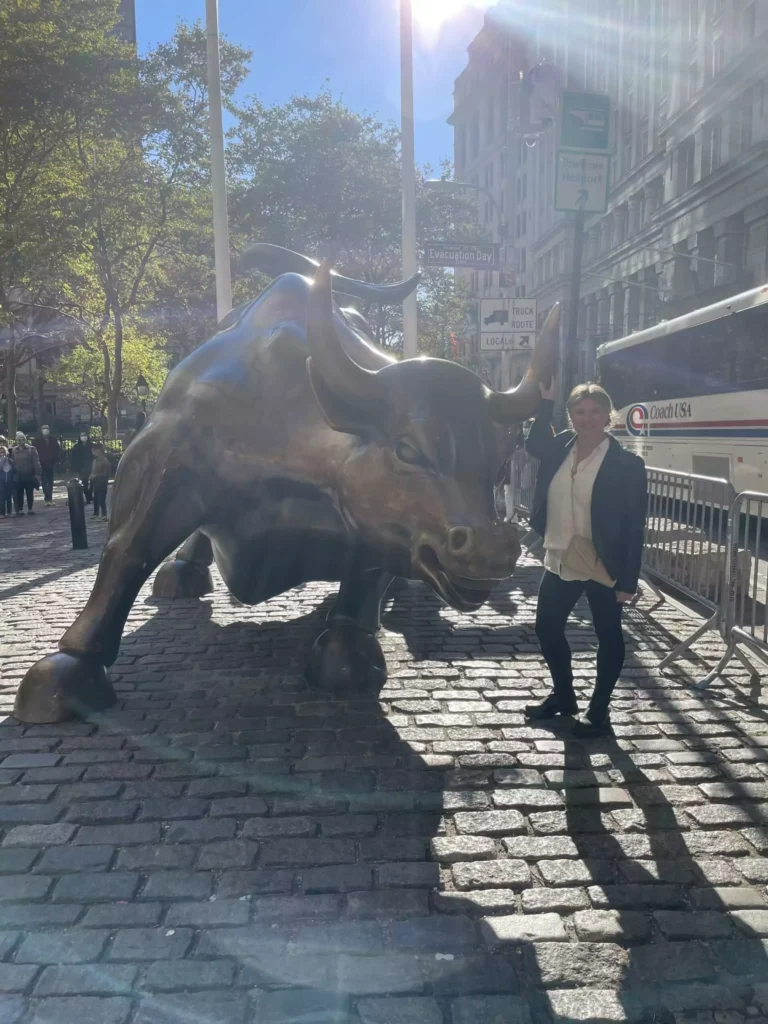 3 Days in New York City - Katie Kinsley standing next to the Charging Bull statue in New York City’s Financial District, smiling beside the iconic bronze sculpture. The cobblestone street and surrounding tourists capture the energy and symbolism of Wall Street.