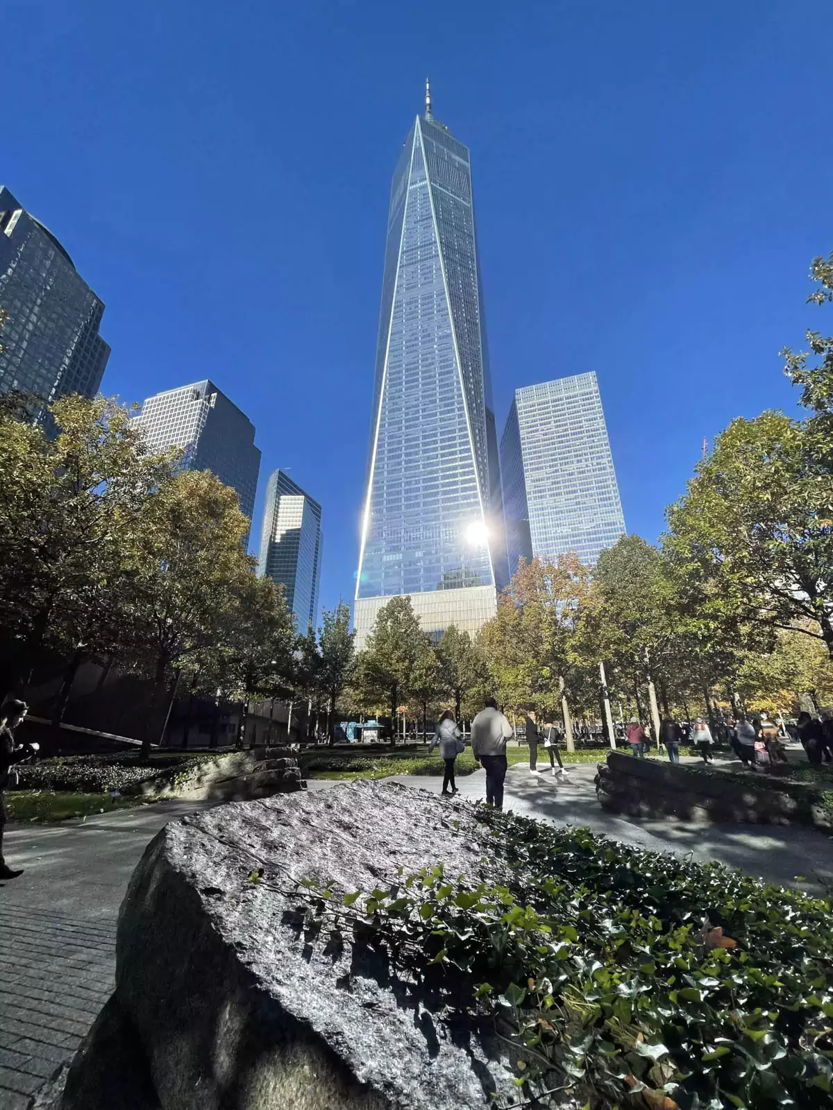 One World Observatory in New York City on a crisp fall day, with the bright sun gleaming off the building’s sleek glass facade. The clear sky and autumn light highlight the tower’s towering presence and reflective surfaces.