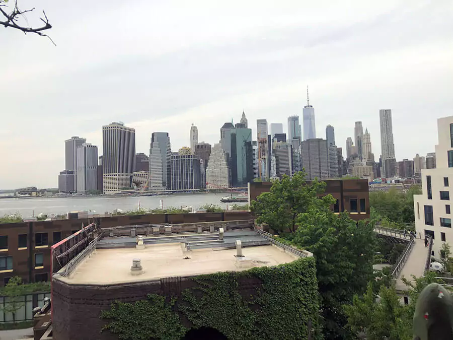 Sweeping view of the Manhattan skyline from Brooklyn, showcasing iconic skyscrapers like the Empire State Building and One World Trade Center across the East River. The foreground includes the waterfront and parts of the Brooklyn Bridge, capturing the classic NYC cityscape.