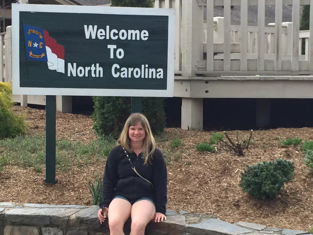 Katie smiling beside a green highway welcome sign that reads ‘Welcome to North Carolina.’ The scene includes roadside greenery and a clear sky, capturing a warm greeting to visitors.