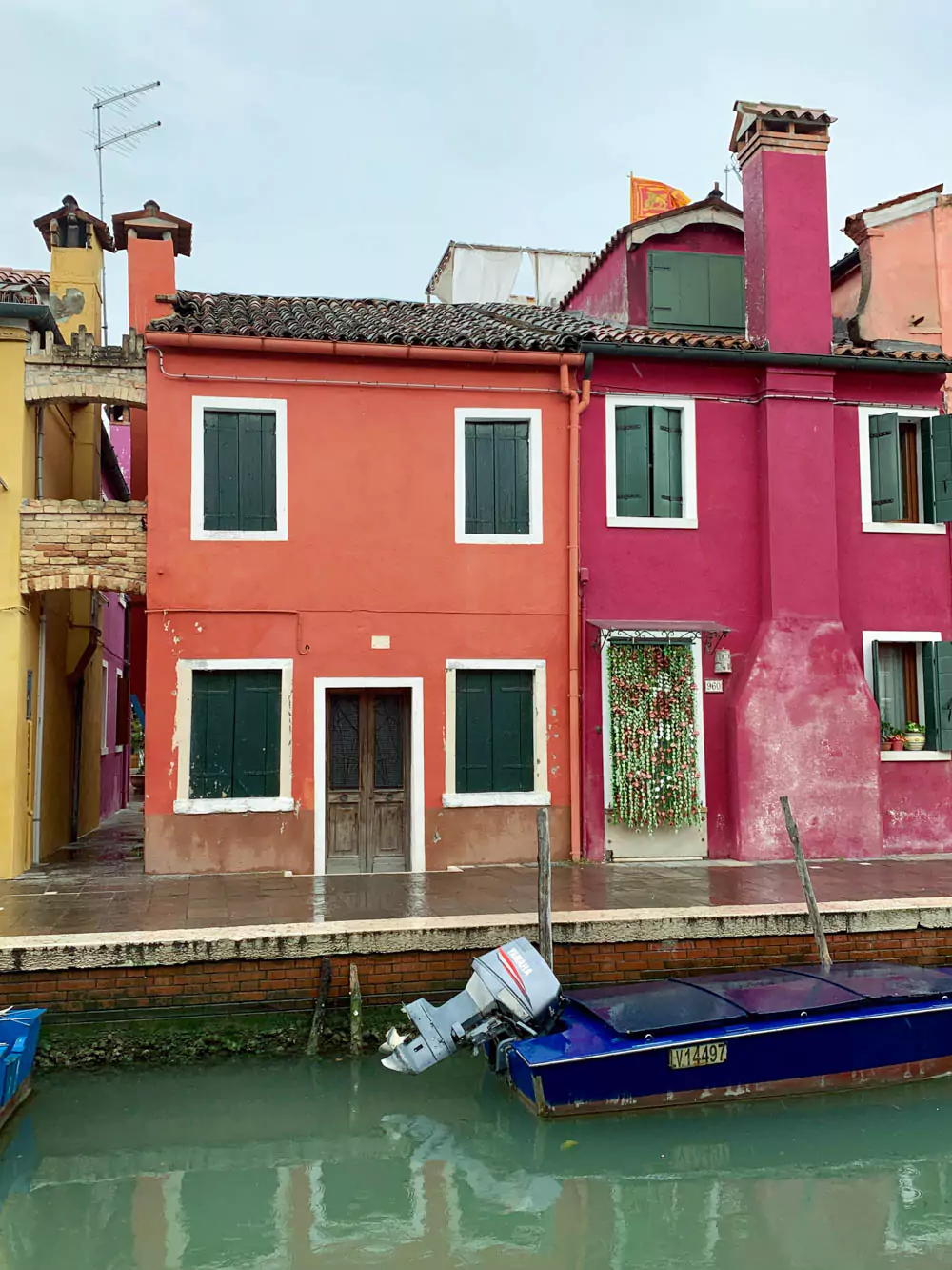 A vibrant orange and pink house sits beside a calm canal in Burano, Italy, with a small blue boat floating in the water out front. The colorful facade and still reflections capture the island’s signature charm and cheerful atmosphere.