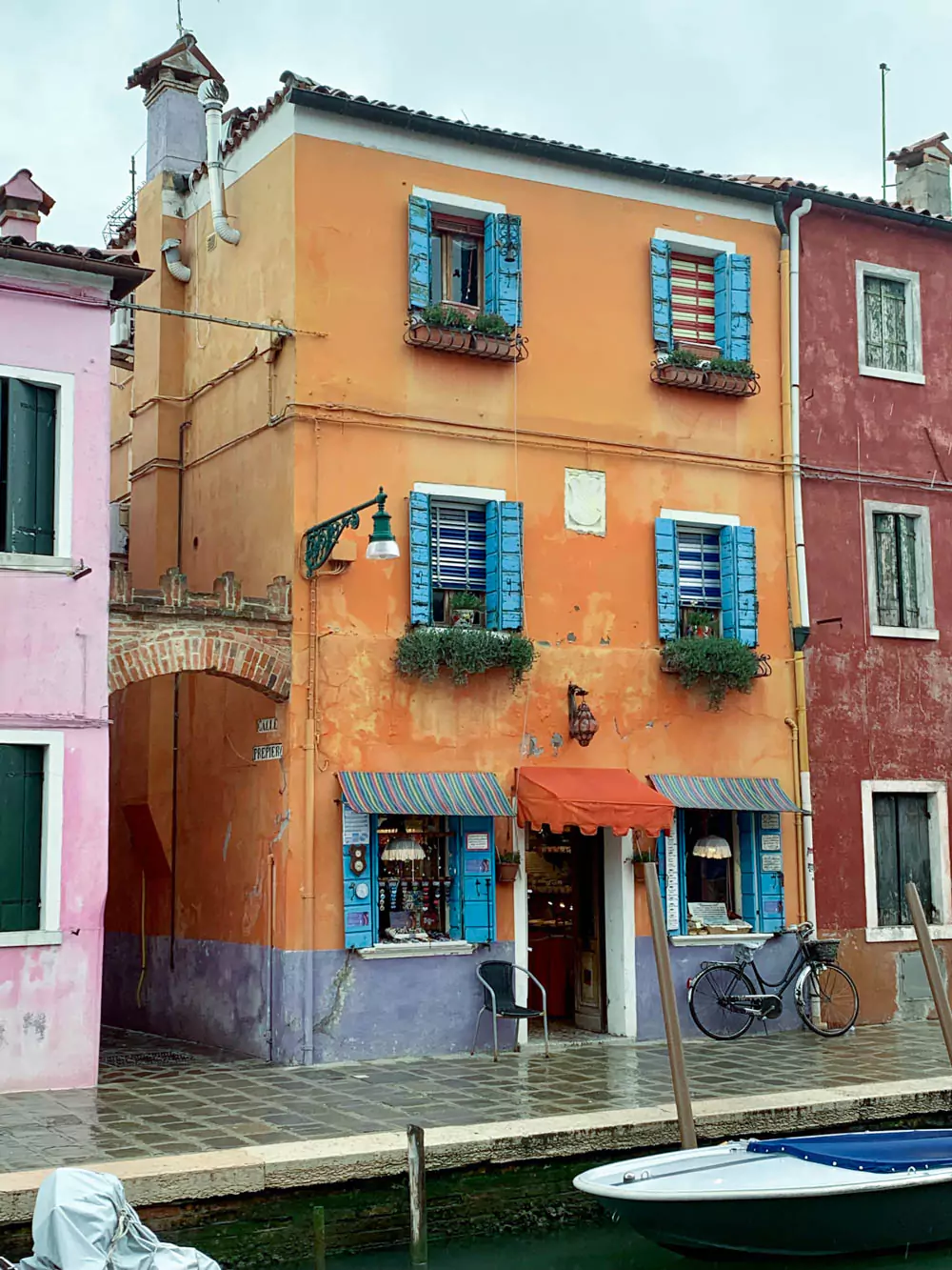 A bright orange building with bold blue shutters stands beside a peaceful canal in Burano, Italy. The striking color combo reflects in the water below, capturing the lively, picturesque vibe the island is known for.