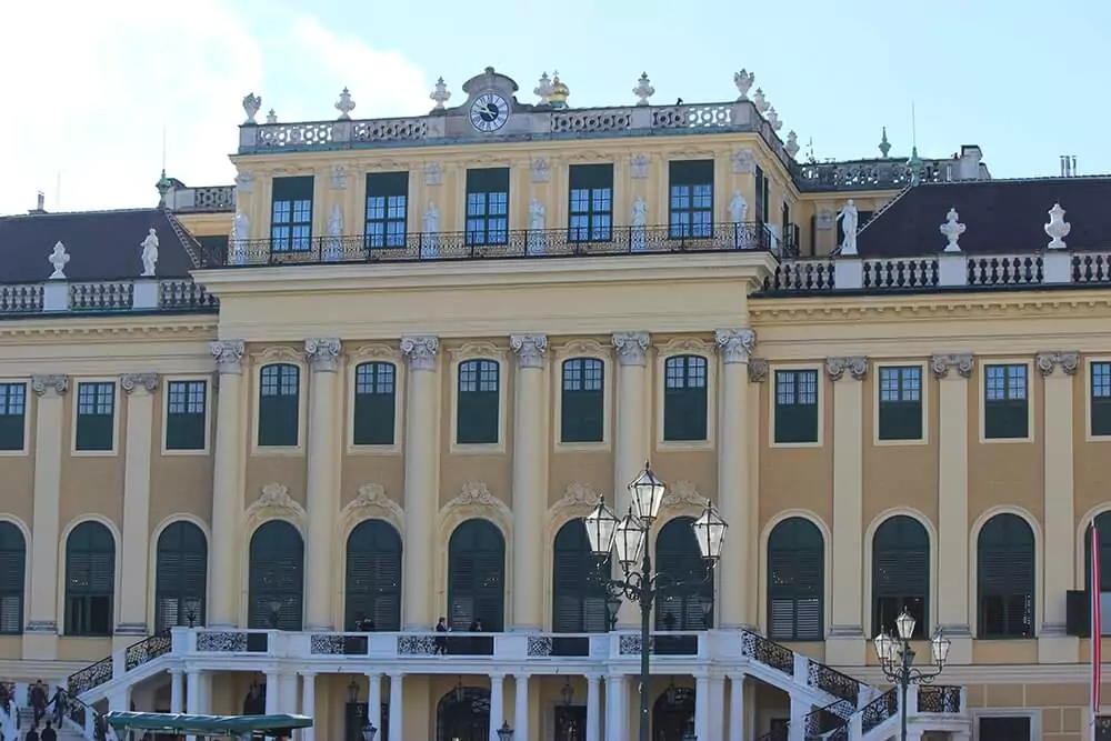 The ornate clock atop Schönbrunn Palace in Vienna features intricate gold detailing, Roman numerals, and a decorative eagle perched above. Set against the palace’s pale yellow facade, the clock adds regal elegance to the historic structure.