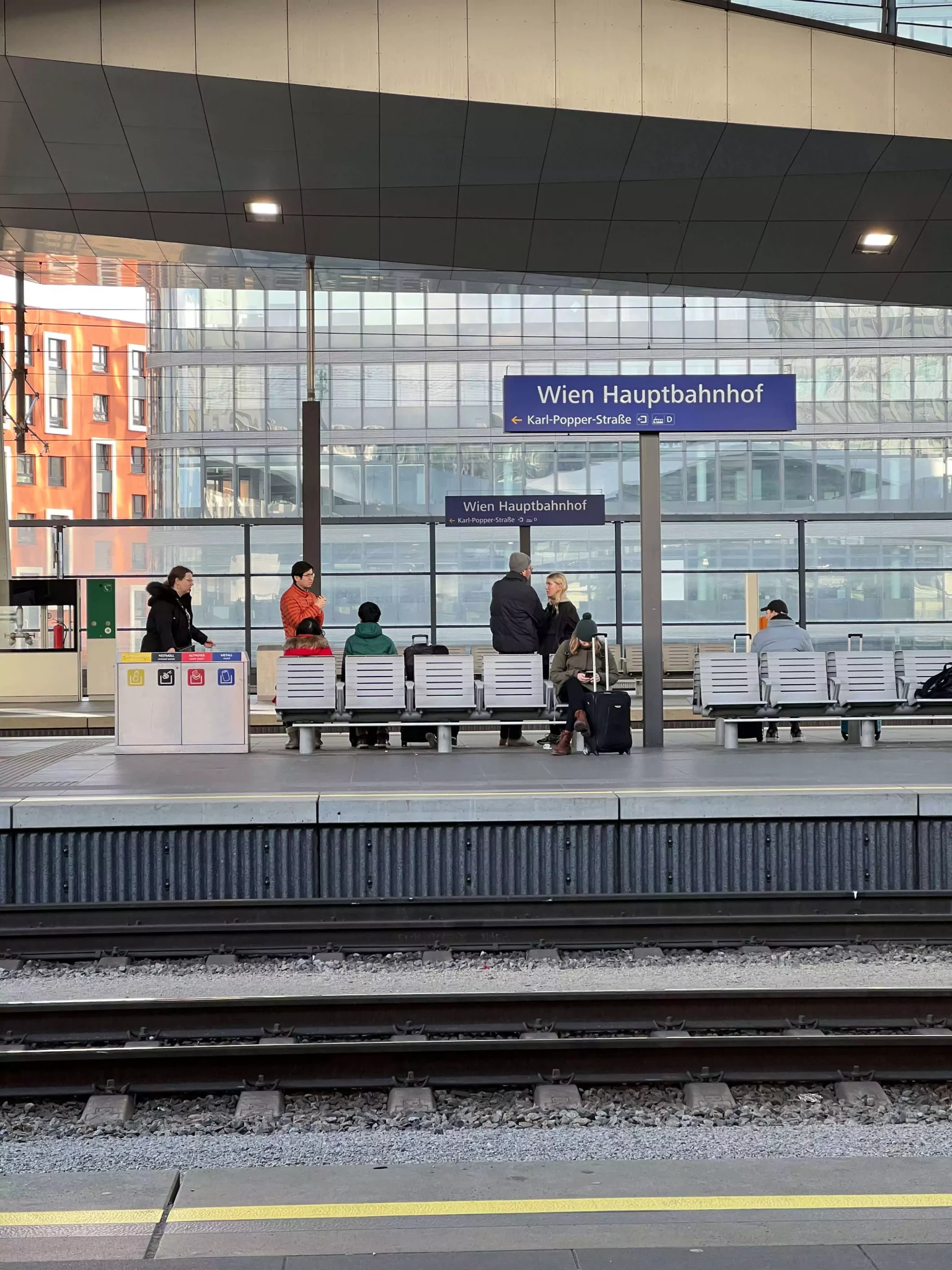 Travelers wait at Wien Hauptbahnhof, Vienna’s main train station, surrounded by modern architecture, digital departure boards, and platform signs. The bustling scene reflects the energy of a major European transit hub.