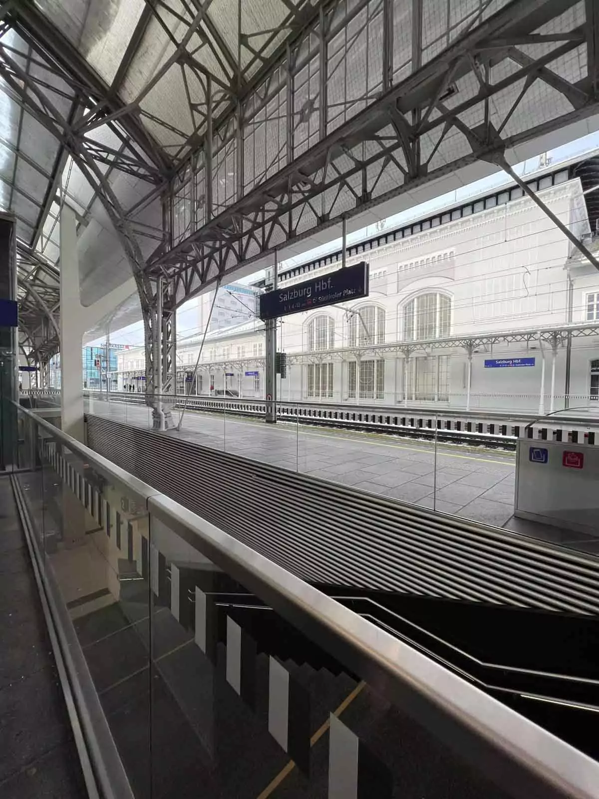 standing on the platform at Salzburg Hauptbahnhof, Austria, with trains arriving in the background, overhead signs, and the station’s modern glass and steel architecture.