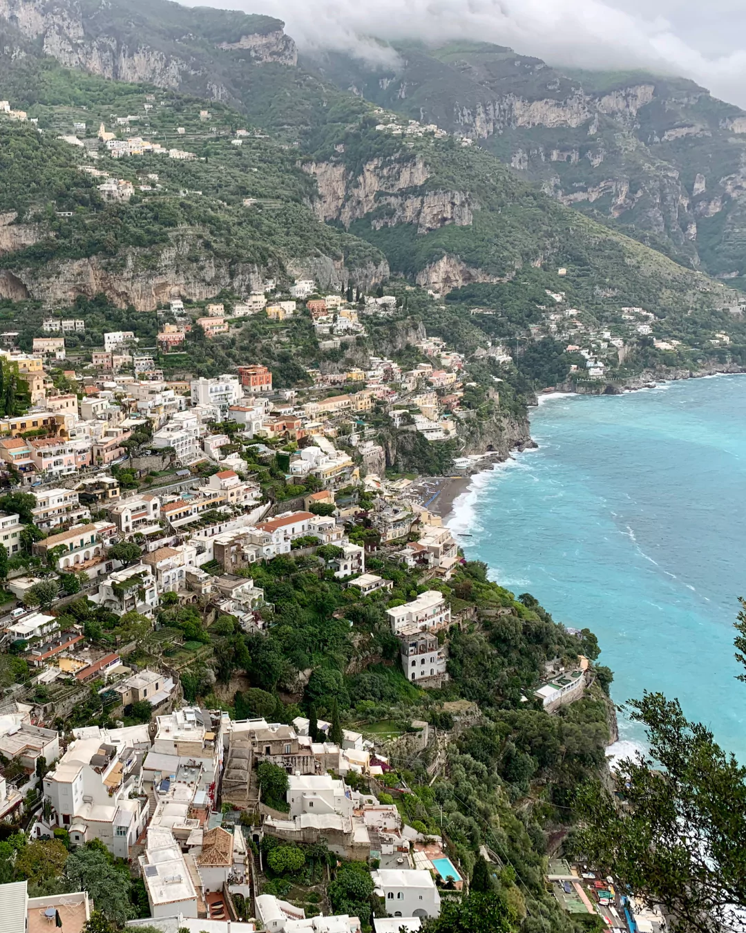 Colorful cliffside buildings of Positano cascading down toward the turquoise waters of the Amalfi Coast in Italy, with terraced homes, lush greenery, and boats dotting the shoreline.