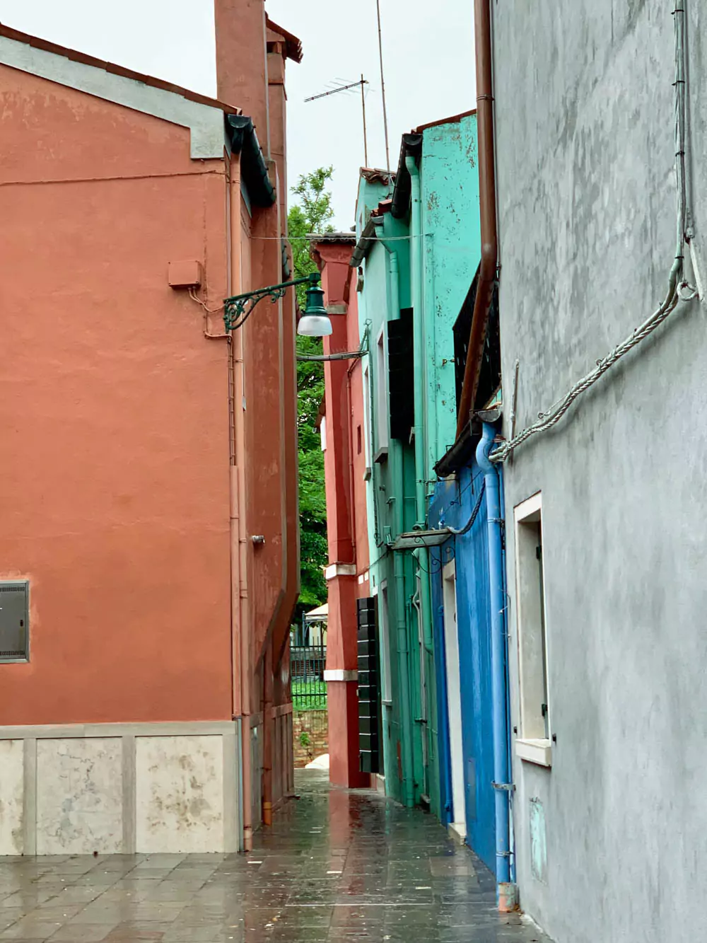 A rainy, narrow alley in Burano, Italy, lined with red, teal, and blue buildings whose wet walls glisten under the overcast sky.