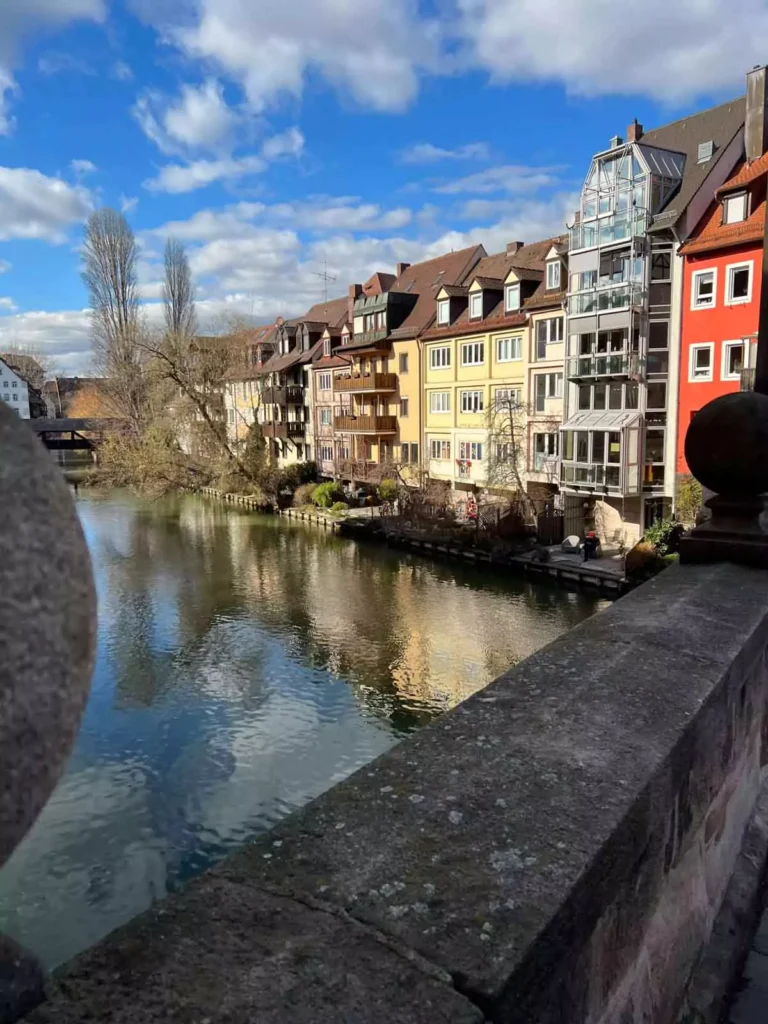 View of the Pegnitz River in Nuremberg, Germany, lined with charming yellow, orange, and red historic buildings reflected in the calm water, capturing the city’s picturesque Old Town atmosphere.