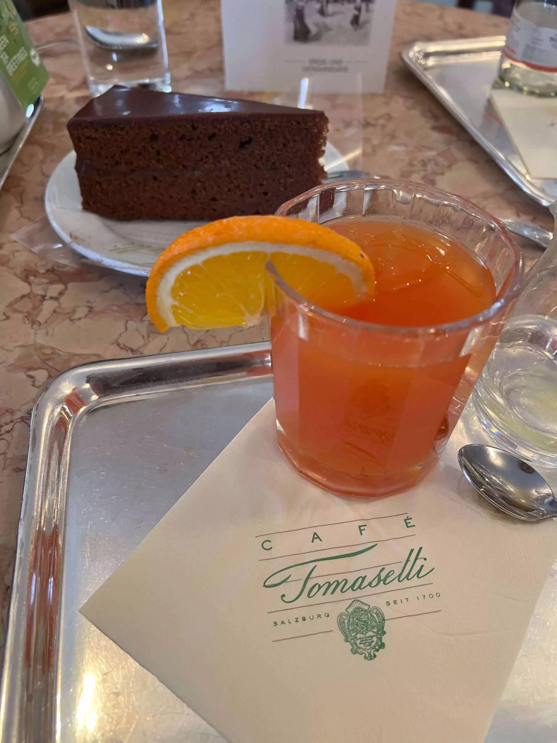 A Campari orange drink at Café Tomaselli in Salzburg, Austria, served in a stemmed glass with ice and a slice of orange. The vibrant color and classic presentation reflect the timeless charm of this historic café.