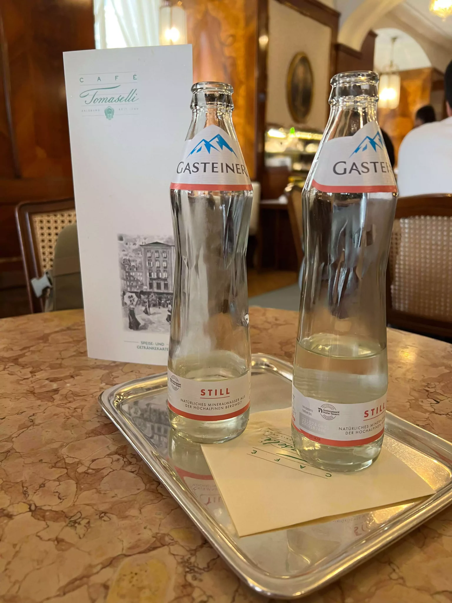 Two glass bottles of still water sit neatly on a table at Café Tomaselli in Salzburg, Austria, alongside a classic printed menu. The setup reflects the café’s timeless elegance and attention to detail in a historic setting.