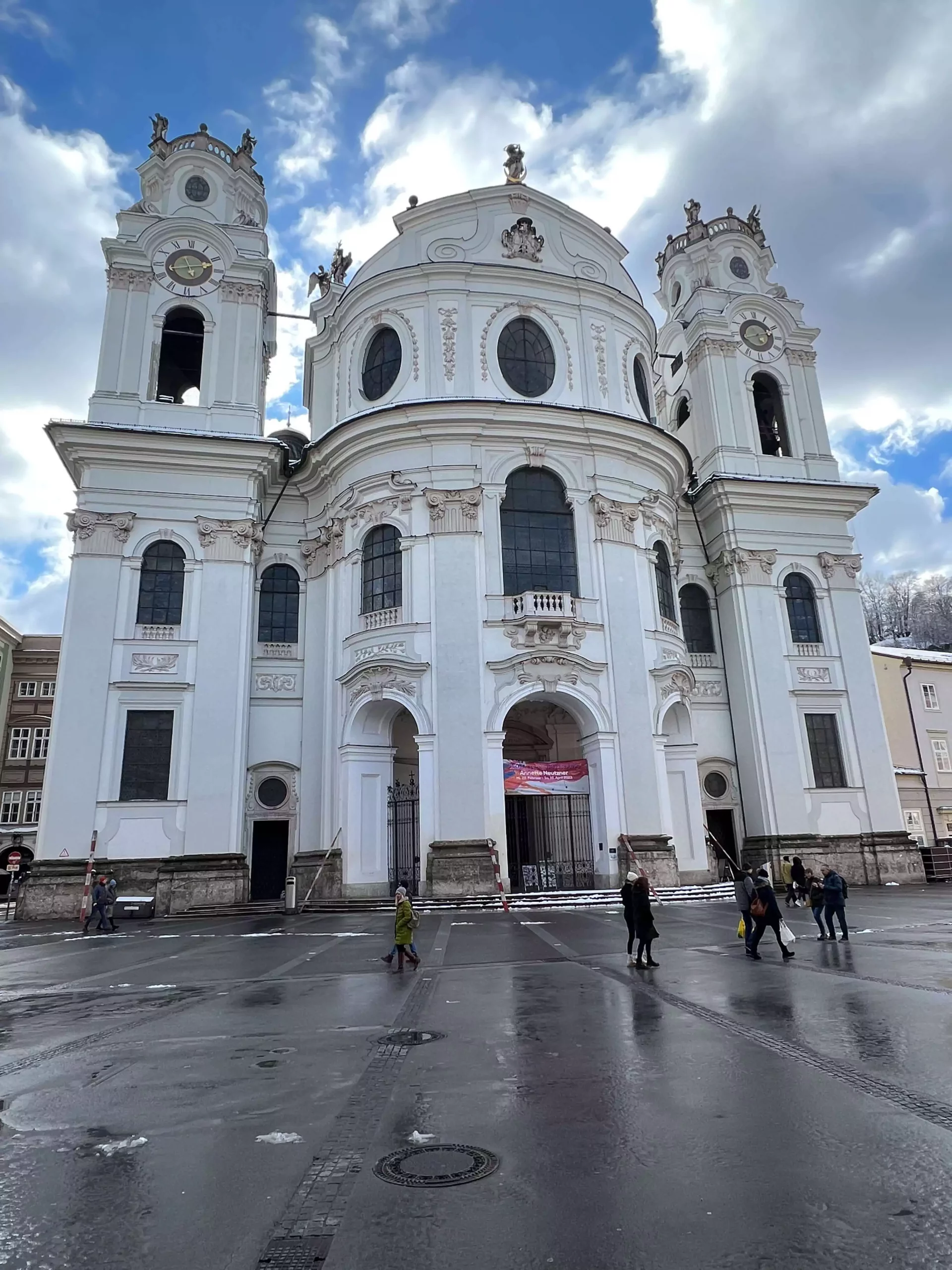 The exterior of Kollegienkirche in Salzburg, Austria, features a grand Baroque facade with soaring columns, curved gables, and detailed stucco work. The church’s pale stone glows in natural light, standing tall against the sky in the heart of the city.