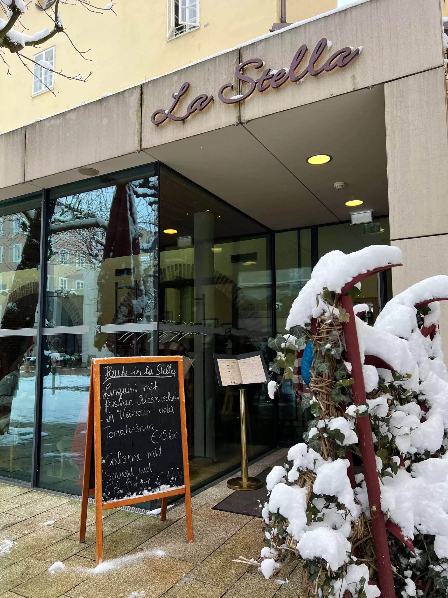 The entrance to La Stella in Salzburg, Austria, set on a sunny street with simple signage and outdoor tables ready for lunch service. The cozy, inviting facade hints at the casual Italian dining experience inside.
