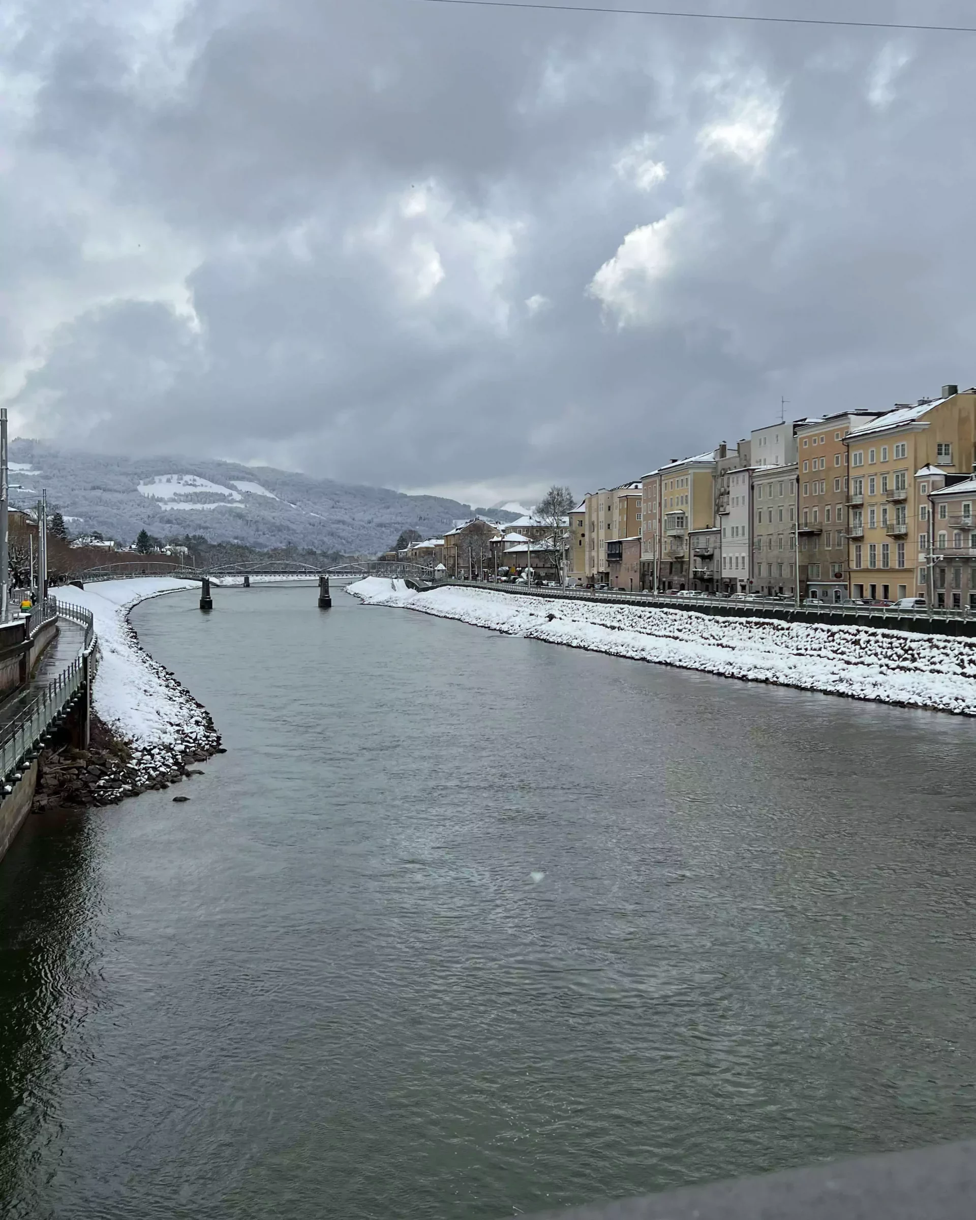 The Salzach River flows calmly through Salzburg, Austria, with snow lining its banks near the Old Town. Historic buildings and spires rise in the distance, adding charm to the wintry riverside view.