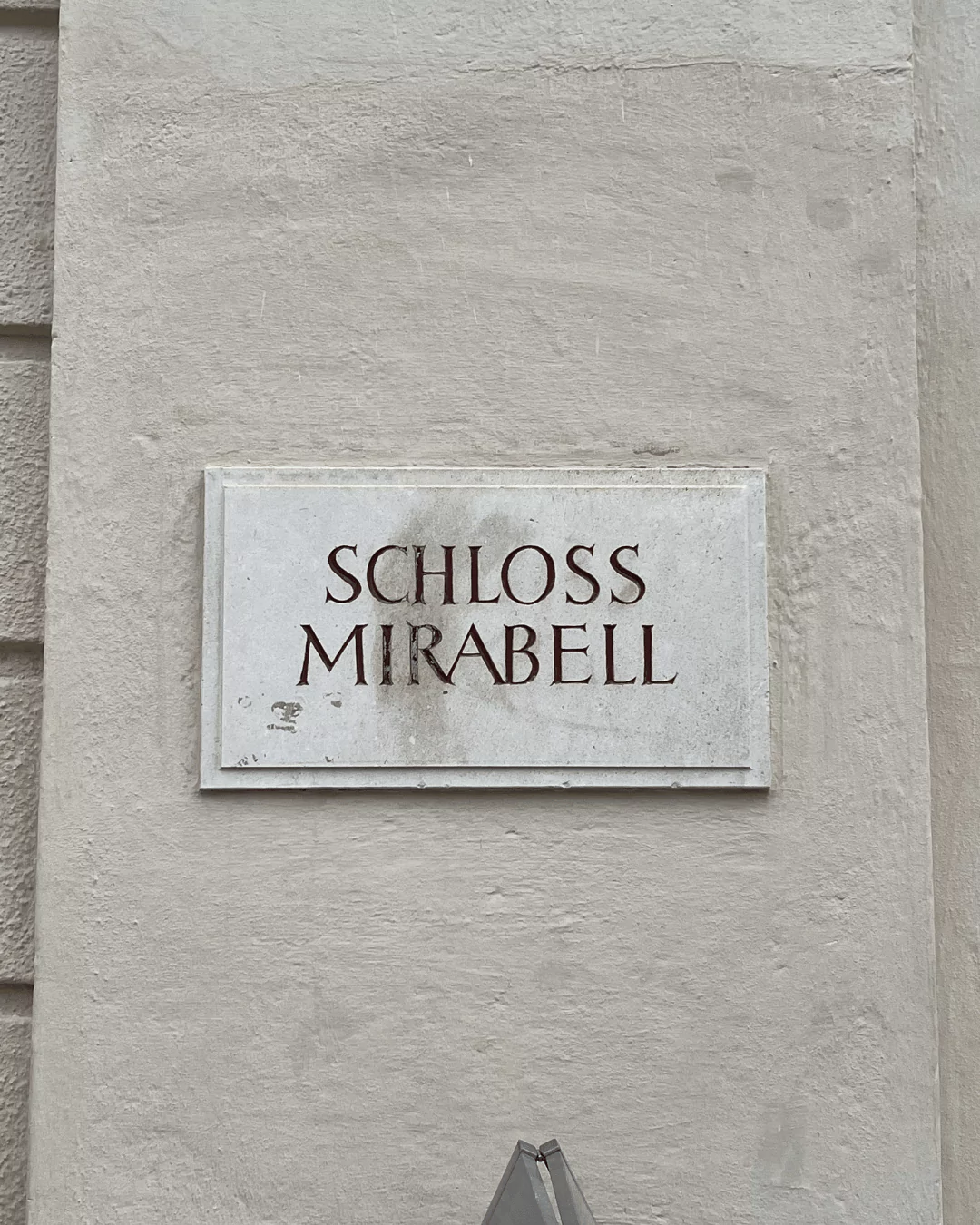 A sign reading “Schloss Mirabell” is mounted on the side of the building, set against the pale stone facade. The simple, elegant lettering marks the entrance to the historic Salzburg landmark.