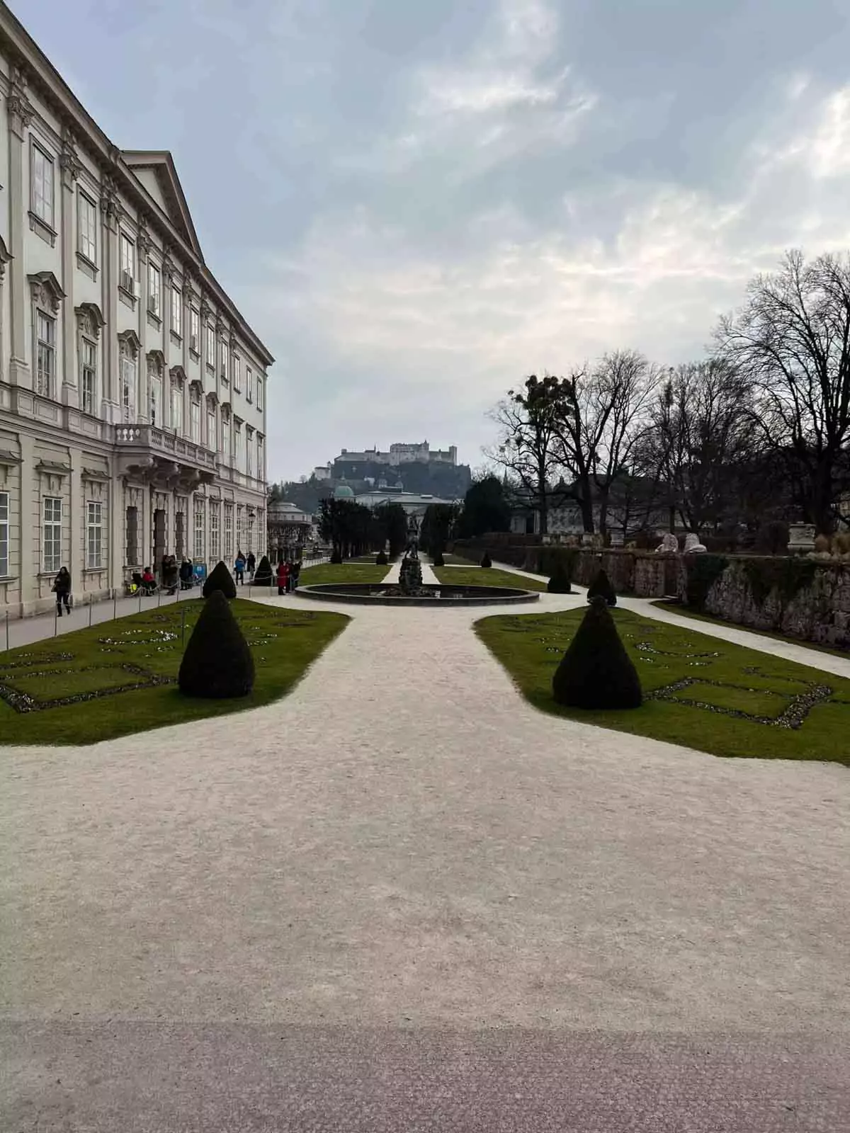 Schloss Mirabell Gardens in Salzburg, Austria, on an overcast day, with trimmed hedges, baroque statues, and flowerbeds set against the cloudy sky and the elegant palace in the background.