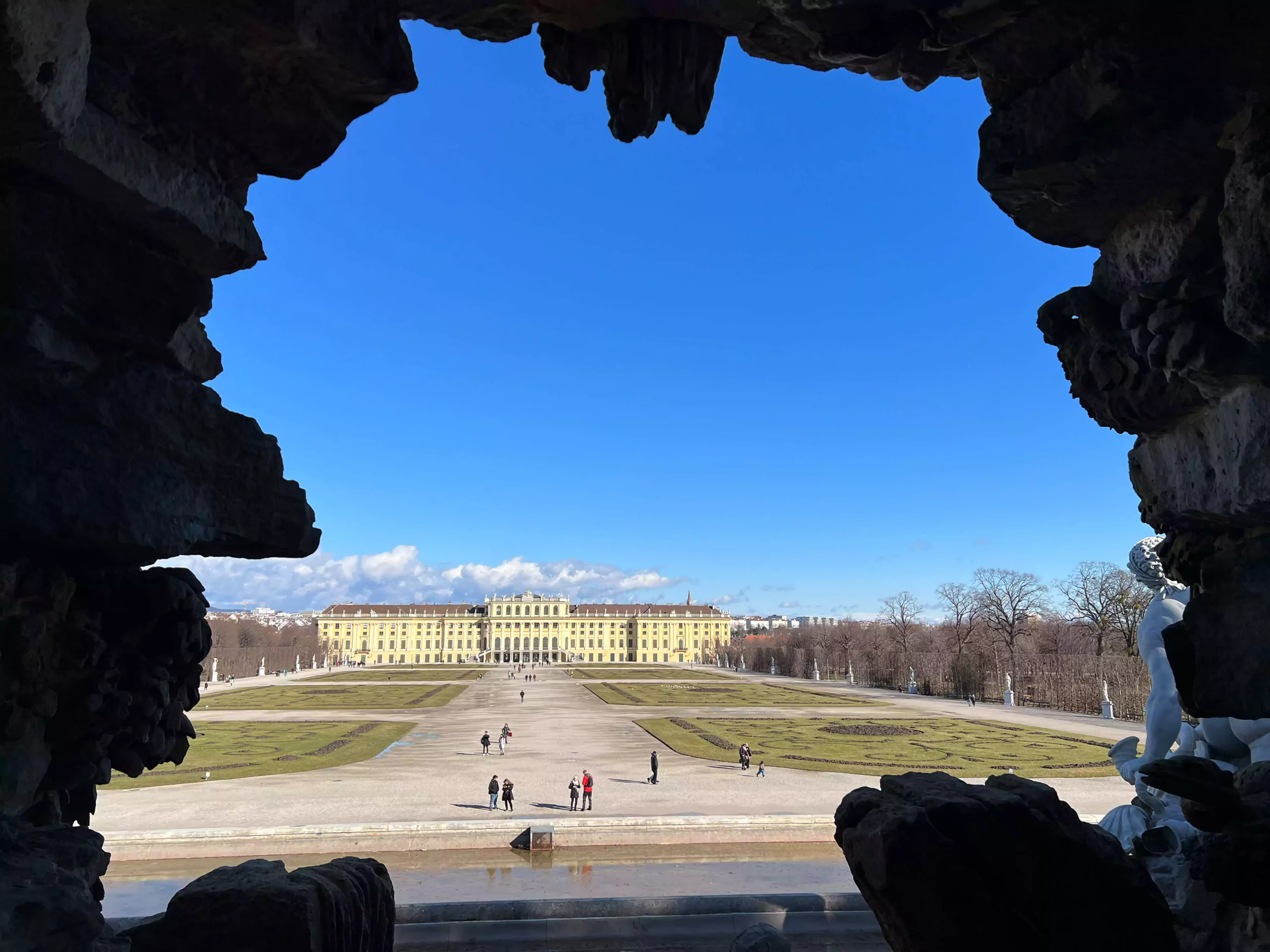 View of the Schönbrunn Palace gardens in Vienna, Austria, from behind the Neptune Fountain, with sculpted figures in the foreground and manicured lawns, floral patterns, and the Baroque palace stretching out in the distance.