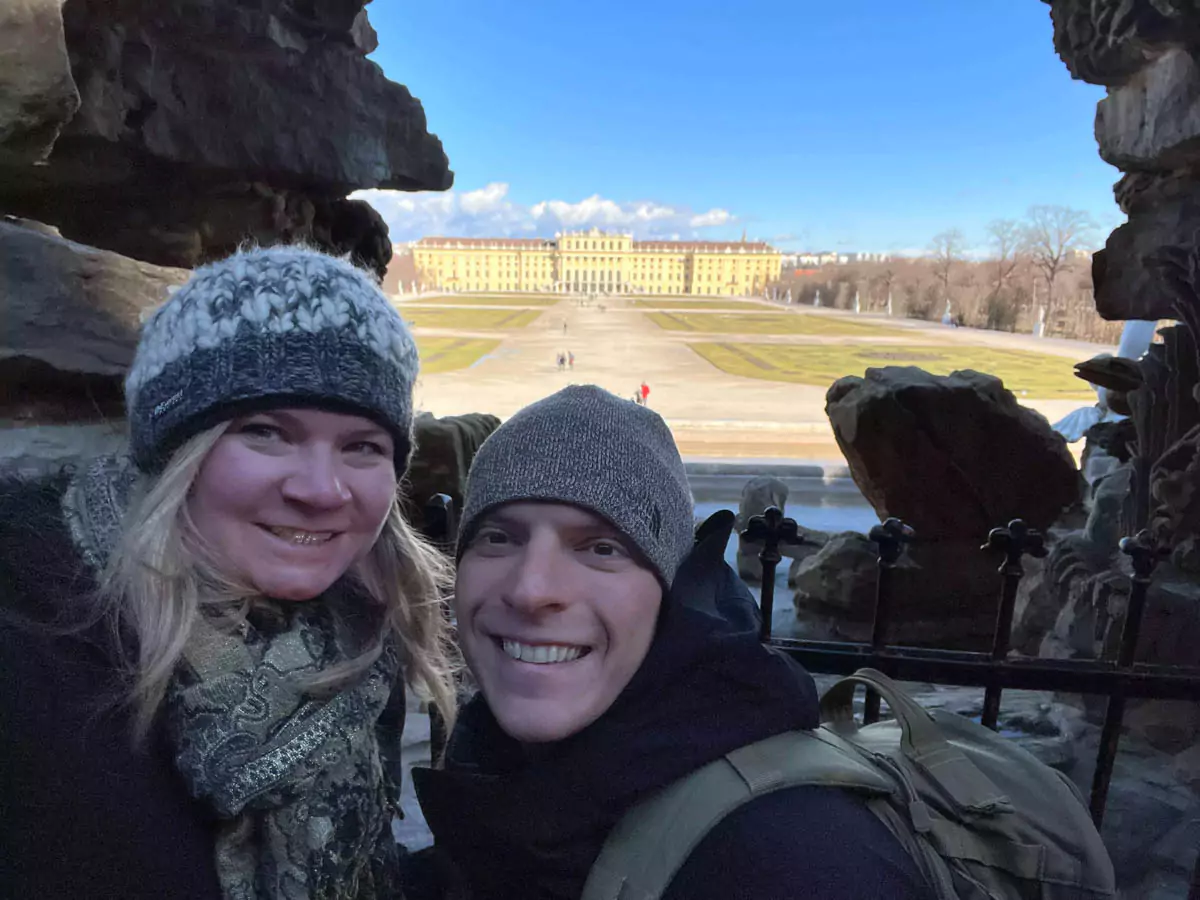 Katie and Zack standing behind the Neptune Fountain at Schönbrunn Palace in Vienna, Austria, with cascading water, classical statues, and the grand palace gardens stretching out in the background.
