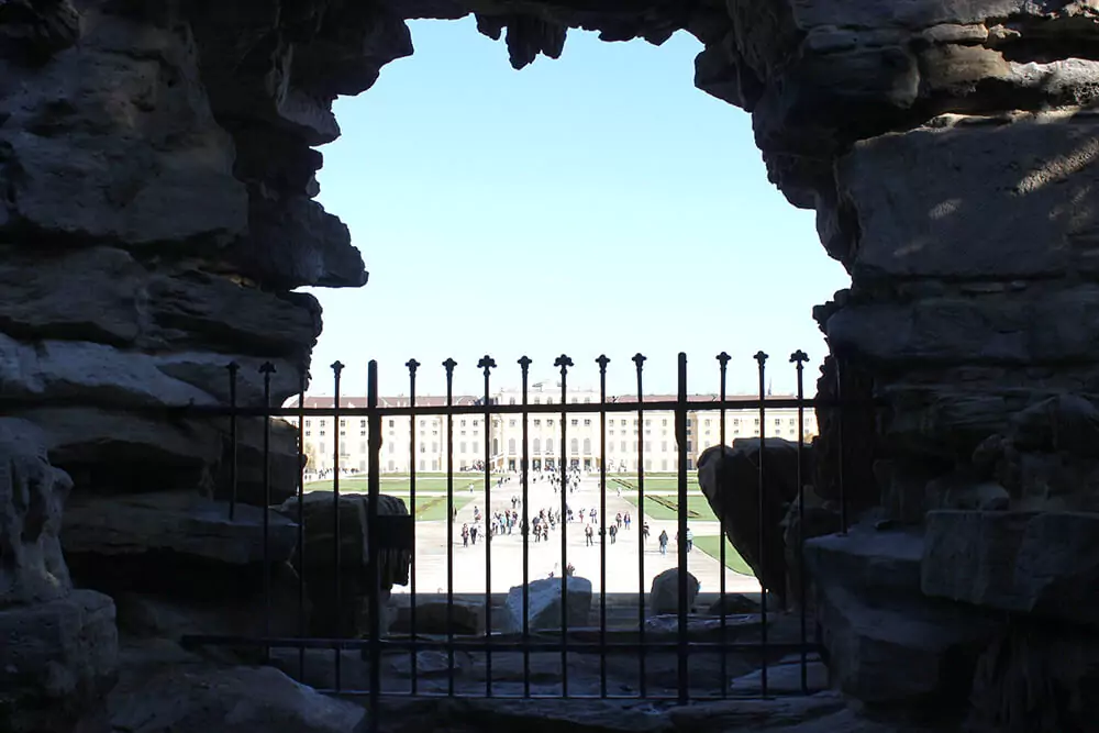 Schönbrunn Palace and its perfectly manicured gardens in Vienna are beautifully framed by the rocky arch of the Neptune Fountain. The stone sculptures and cascading water in the foreground create a dramatic natural frame for the grand imperial view.