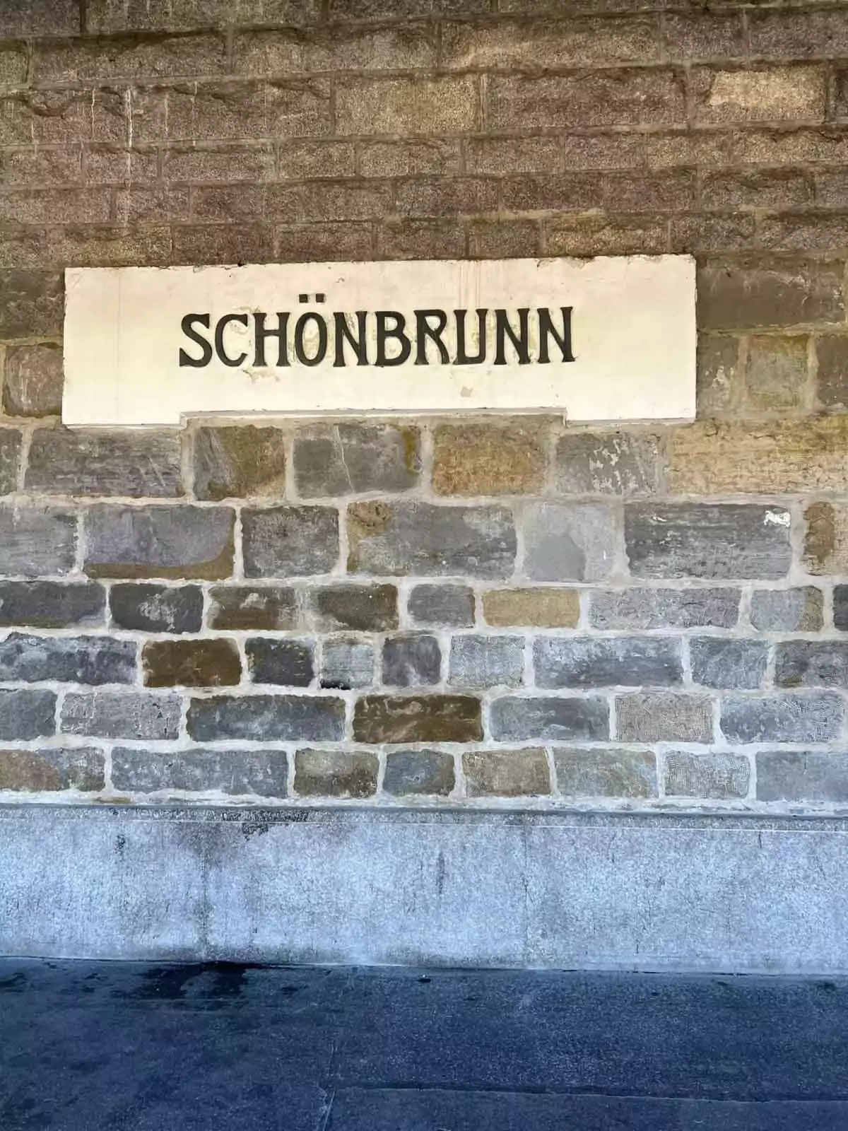 Schönbrunn Station sign embedded in a stone wall in Vienna, Austria, with classic black lettering on a white background, marking the U-Bahn stop near Schönbrunn Palace.