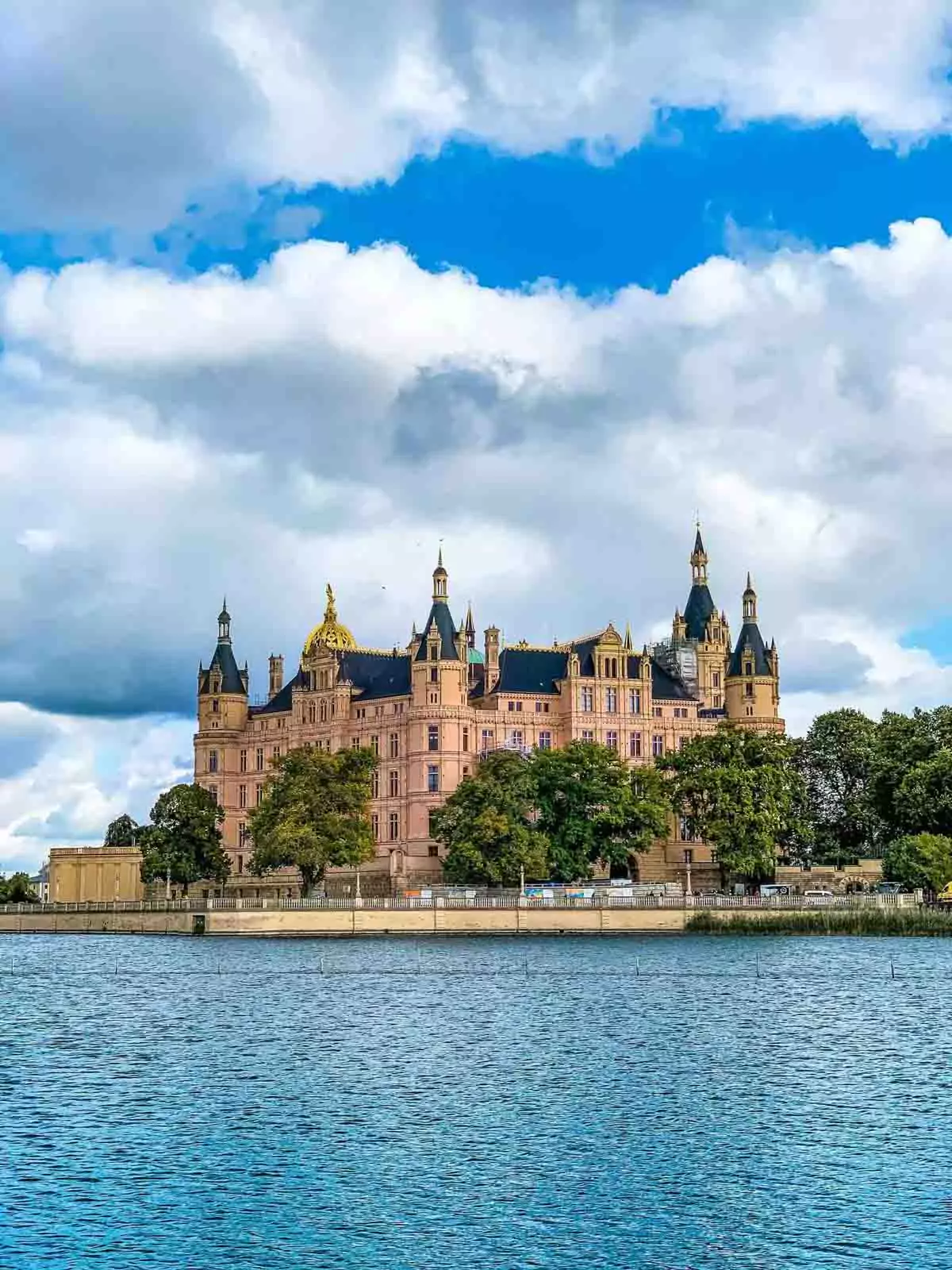 Fairytale-like Schwerin Castle in Germany, seen from across the water, with its ornate towers and golden dome reflected in the lake, surrounded by lush greenery and historic charm.