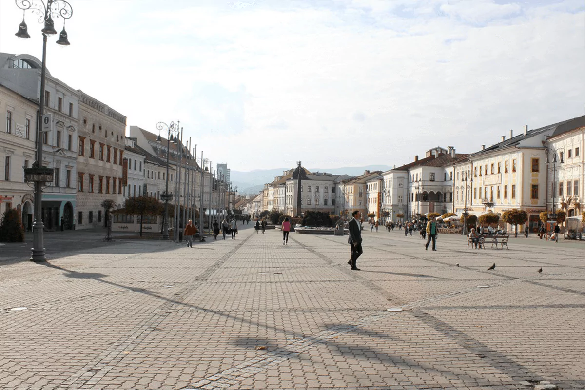 SNP Platz in Banská Bystrica, Slovakia, a historic town square lined with pastel-colored buildings, cobblestone walkways, and the central Marian column, with locals and tourists enjoying the open space 
