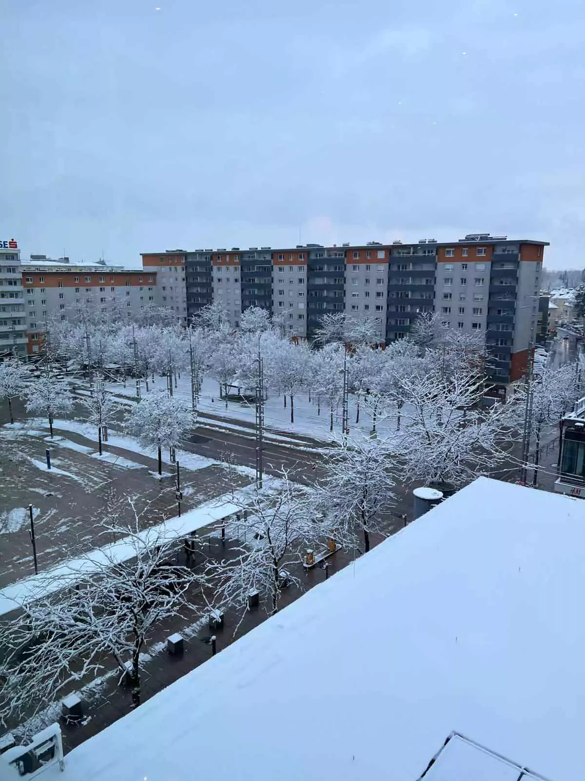 Snowy morning scene outside the Salzburg train station in Austria, with light snow covering the ground, people bundled in winter coats, and trains or trams arriving against a wintry urban backdrop.