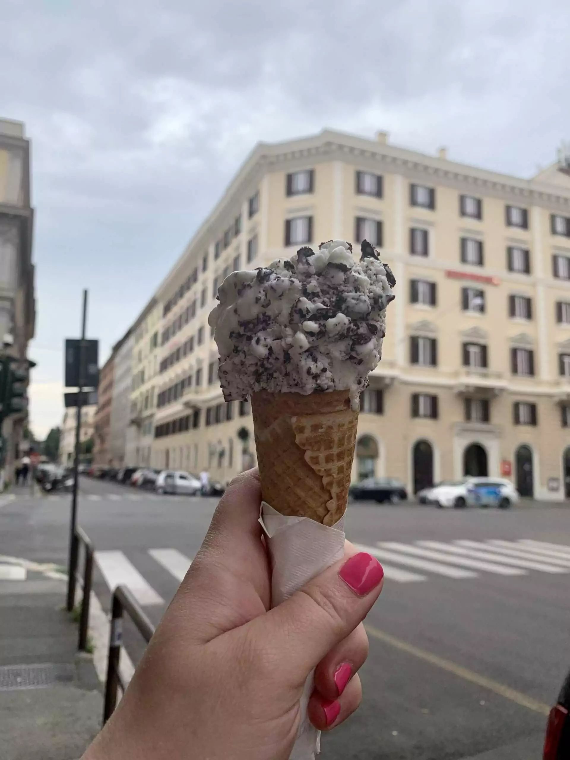 Katie’s hand holding a cone of stracciatella gelato in Rome, with creamy vanilla ice cream swirled with chocolate flakes, captured against a charming city street backdrop.
