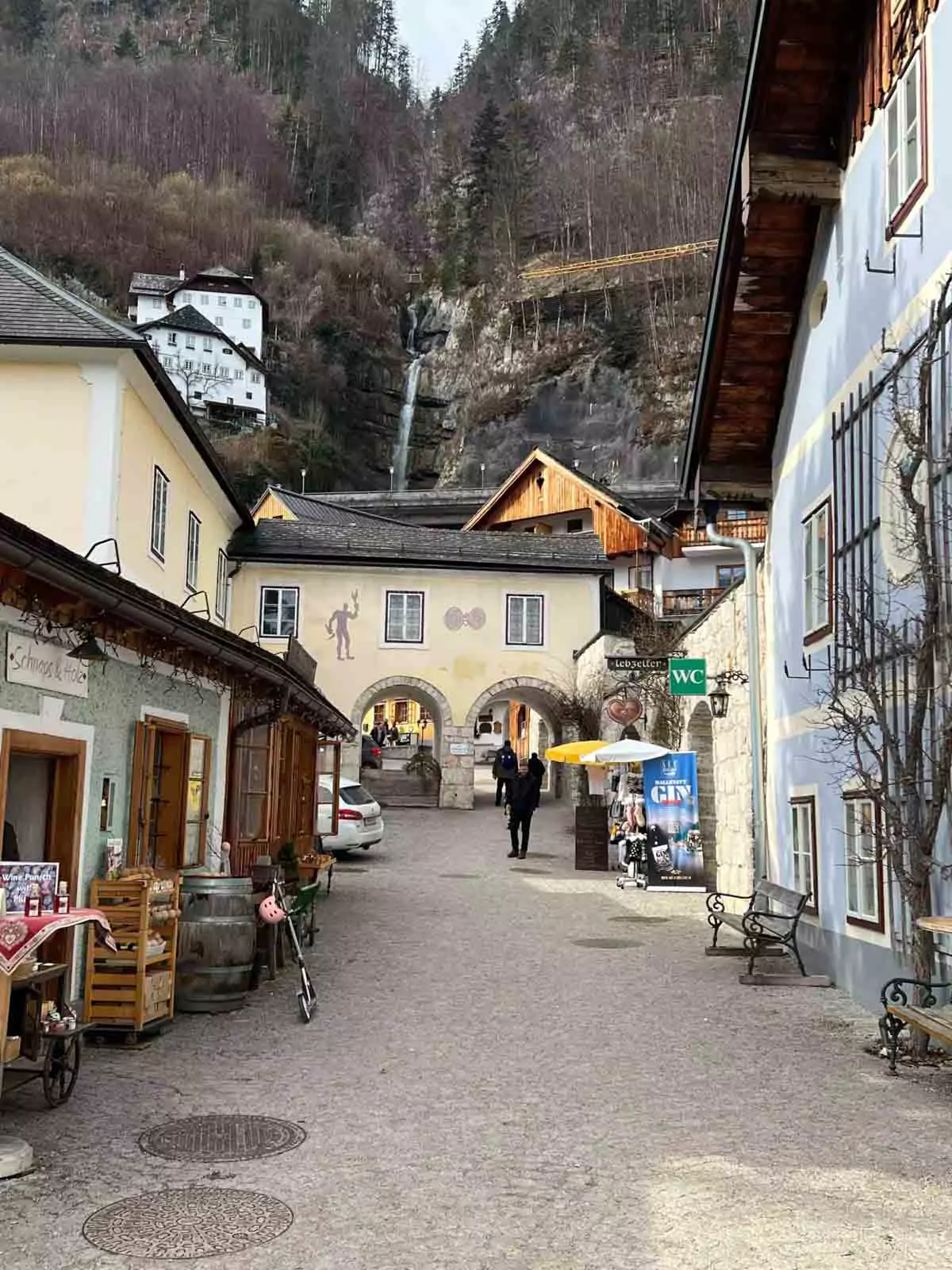 Picturesque street view in Hallstatt, Austria, lined with traditional alpine houses and shops, with a cascading waterfall visible in the background against the lush mountain slope.