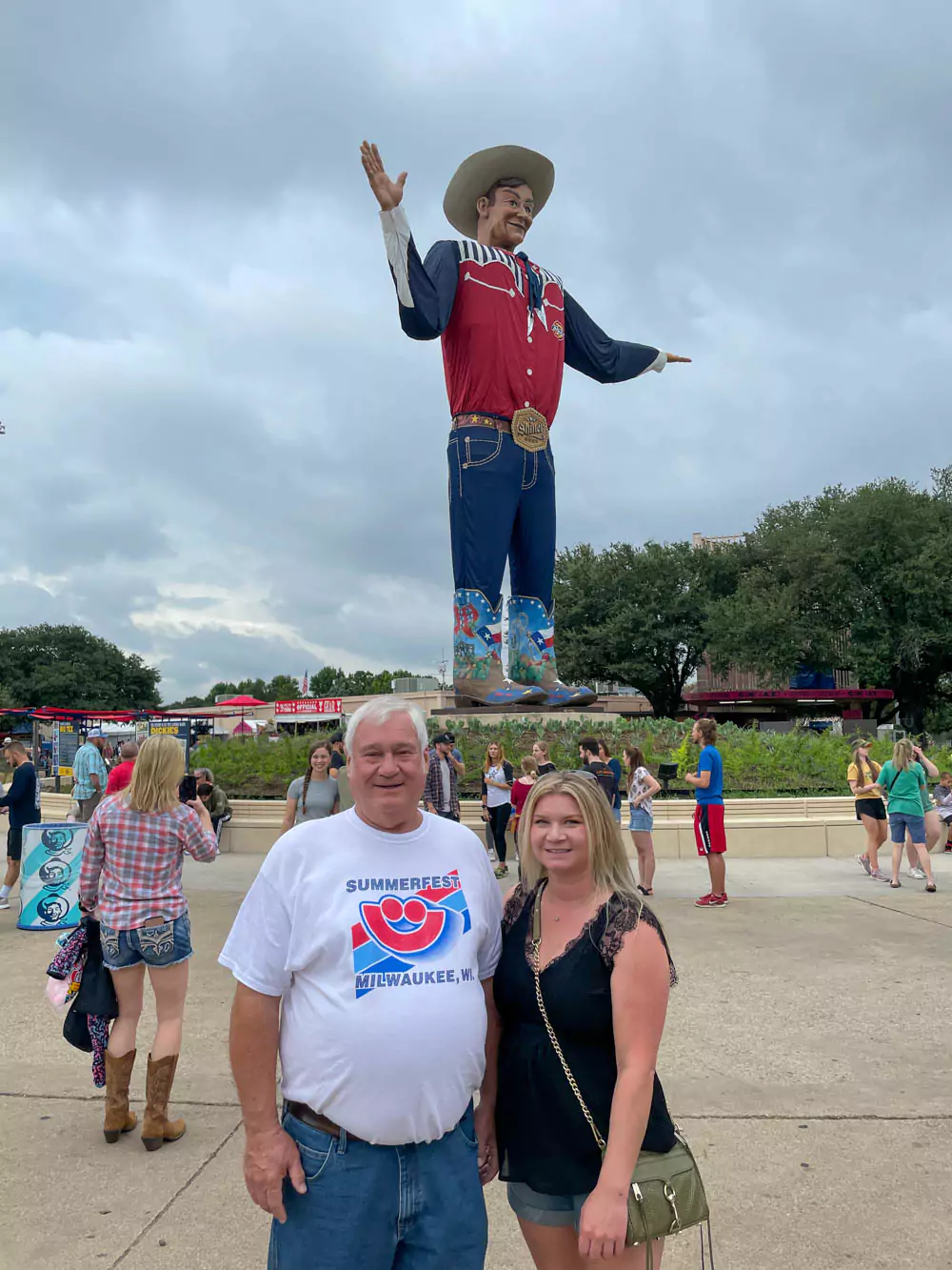 Katie and her dad smile standing beside Big Tex, the towering iconic cowboy statue at the Texas State Fair. The festive fairground atmosphere includes bright colors and clear skies.