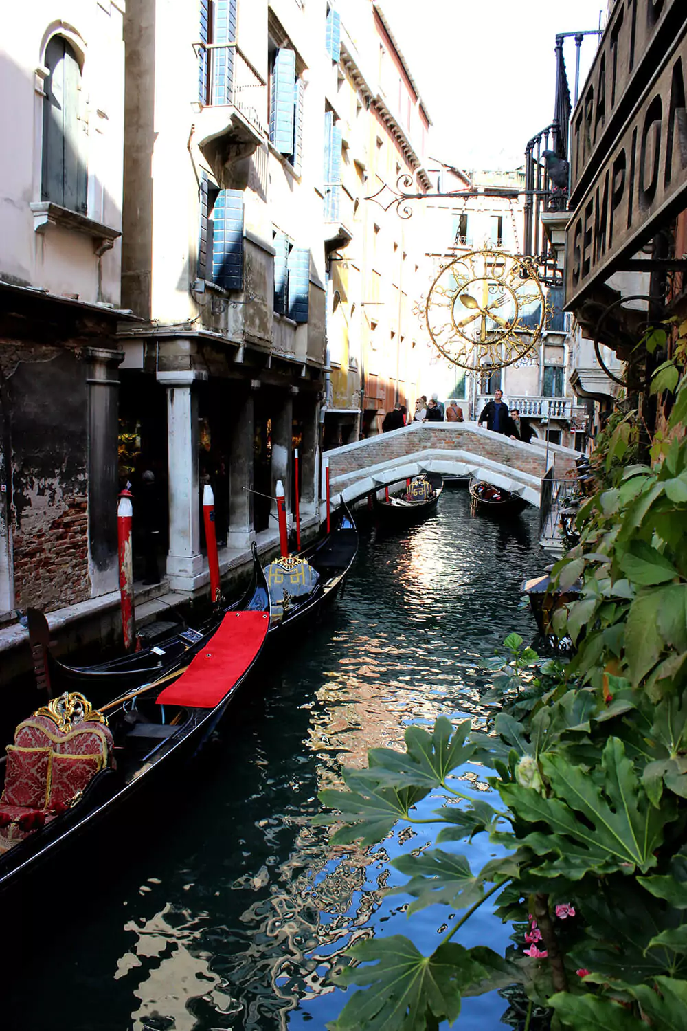 Several black Venetian gondolas float in a calm canal beneath a stone bridge crowded with people. The scene captures the classic charm and bustling activity of Venice’s waterways.