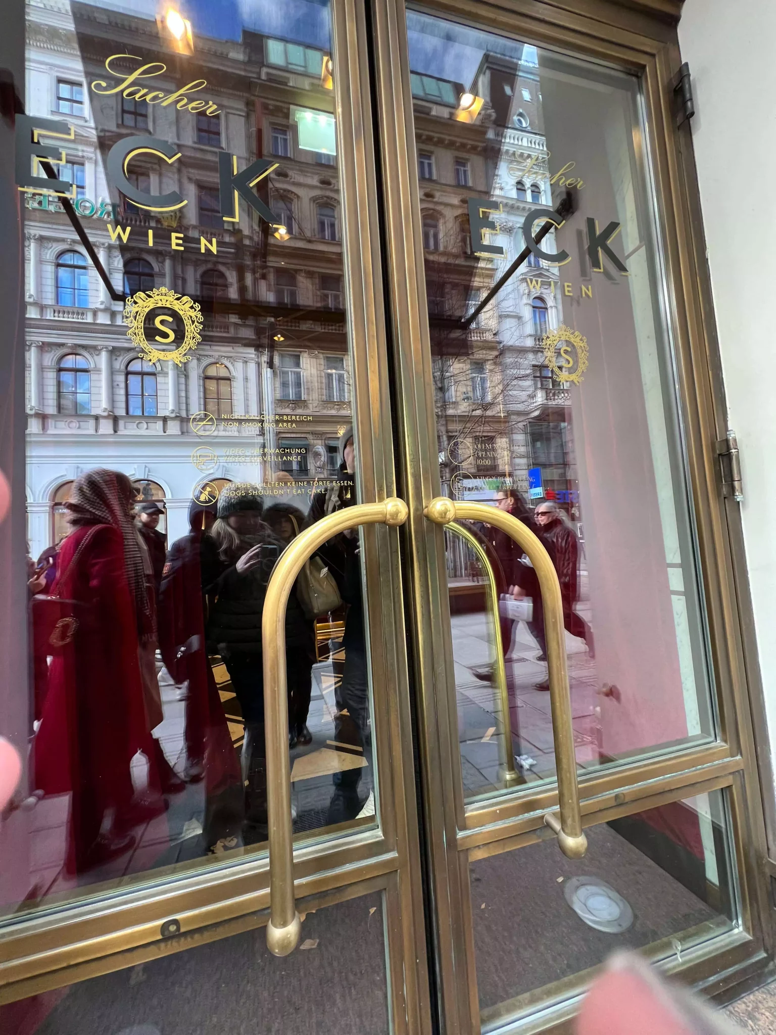 The entrance doors to Eck at Café Sacher in Vienna, Austria, feature dark wood and glass panels with gold lettering, exuding classic elegance. The refined entrance hints at the sophisticated interior of this iconic Viennese establishment.