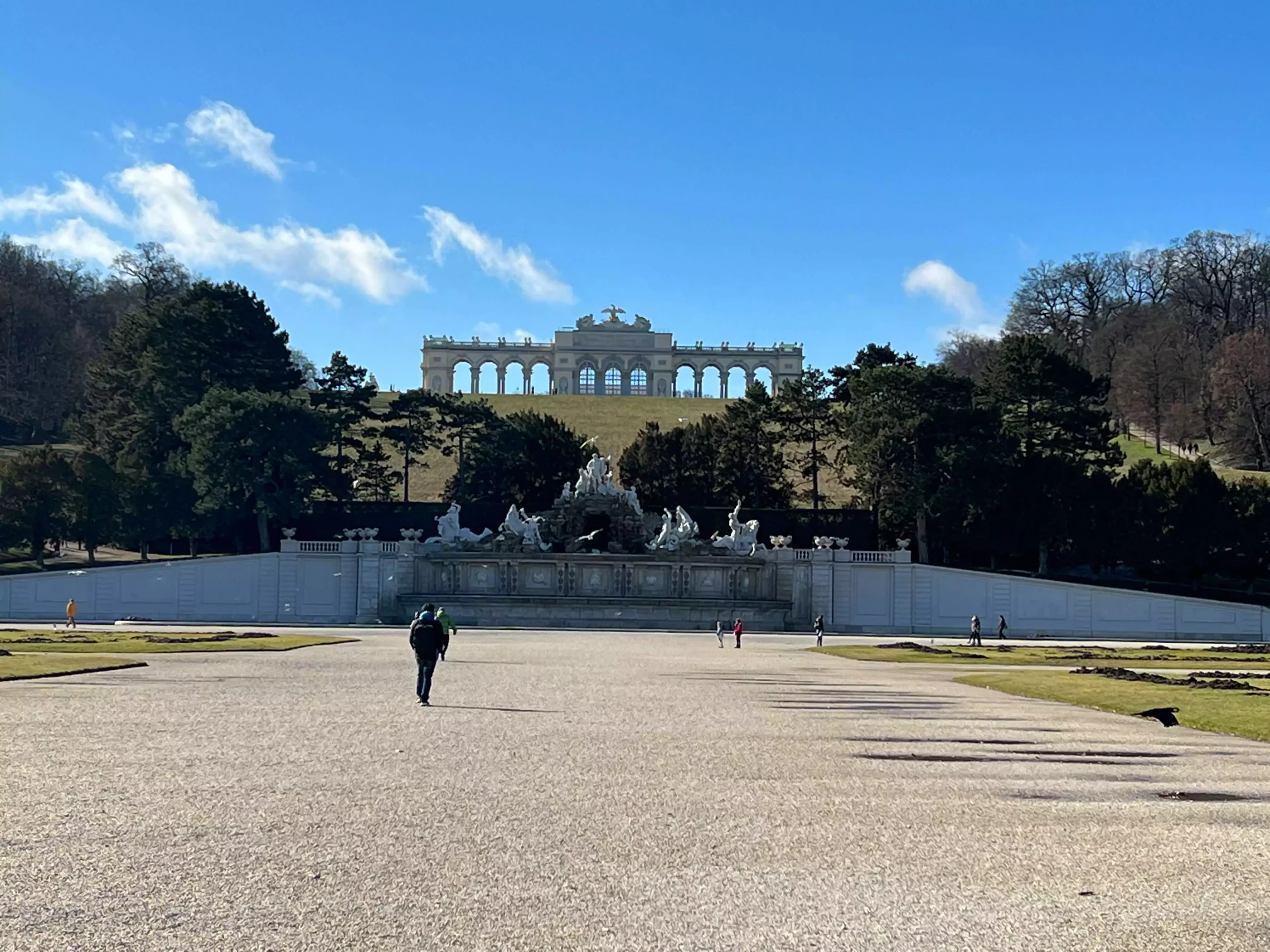 The Gloriette standing atop Schönbrunn Hill in the background, with the Neptune Fountain in the foreground featuring dramatic baroque statues and cascading water, set within the grand gardens of Schönbrunn Palace in Vienna, Austria.