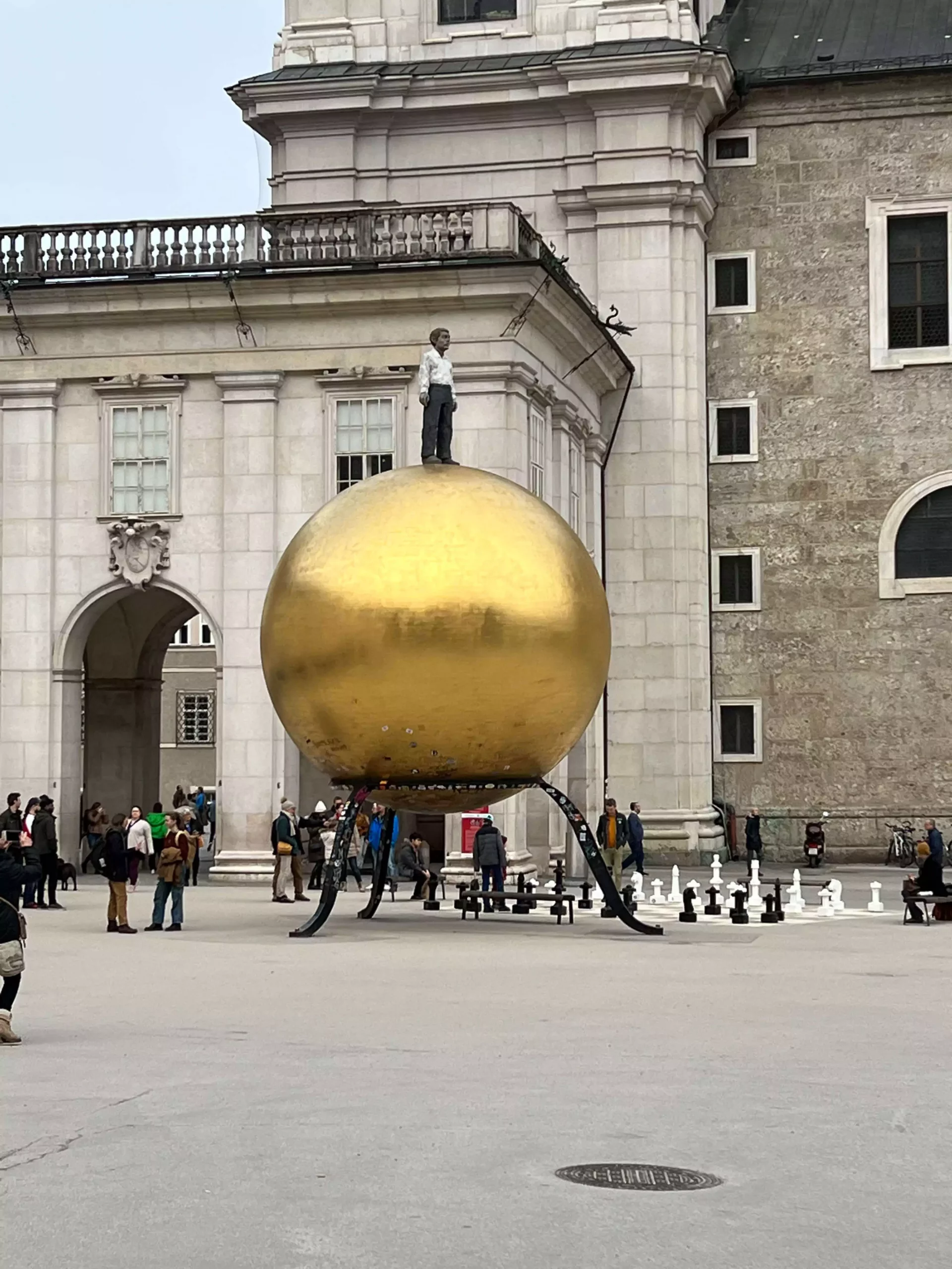 The Goldene Kugel in Salzburg’s Old Town features a large golden sphere sculpture with a standing male figure on top, set against the historic buildings of Kapitelplatz. The modern artwork contrasts playfully with the surrounding baroque architecture.