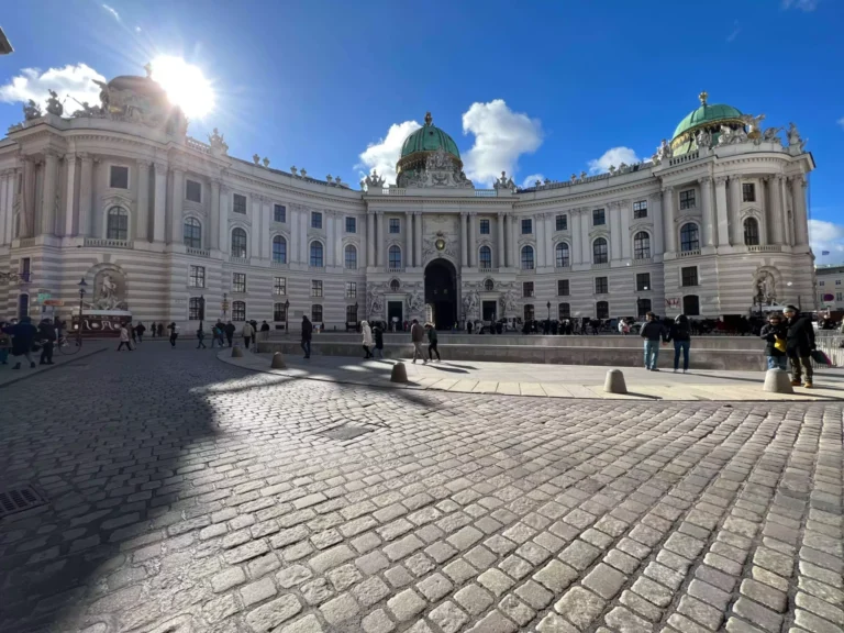 The Hofburg Palace in Michaelerplatz, Vienna, Austria, stands majestically under a clear blue sky, with its grand Baroque façade, domed roof, and ornate detailing gleaming in the sunlight. The historic square buzzes with visitors admiring the imperial architecture.
