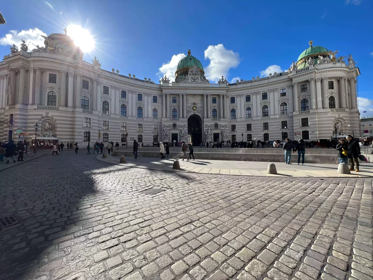 The Hofburg Palace in Michaelerplatz, Vienna, Austria, stands majestically under a clear blue sky, with its grand Baroque façade, domed roof, and ornate detailing gleaming in the sunlight. The historic square buzzes with visitors admiring the imperial architecture.