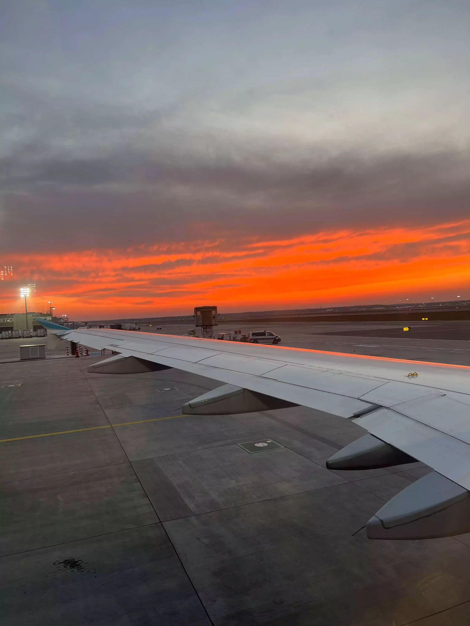 A red, cloudy sunrise glows through the plane window just before takeoff in Germany. The vibrant hues light up the horizon, casting a warm glow over the wing and runway below.