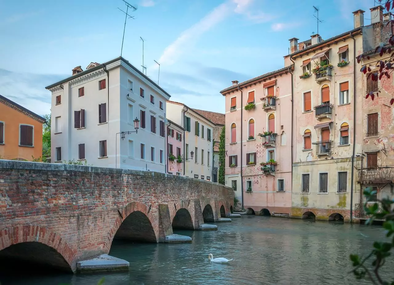 Stone bridge over a calm canal in Treviso, Veneto, Italy, with colorful historic buildings lining the waterway and reflections shimmering beneath the arch on a quiet day.