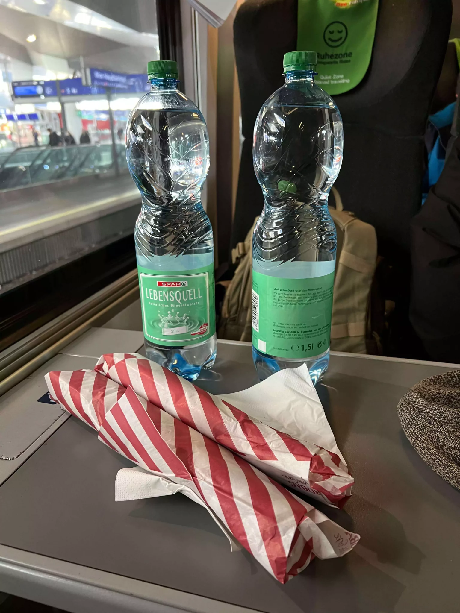 Two wrapped sandwiches and bottles of still water set out for dinner aboard a Vienna-bound train. The simple, on-the-go meal rests on a fold-down tray, capturing the cozy practicality of dining while traveling through Europe.
