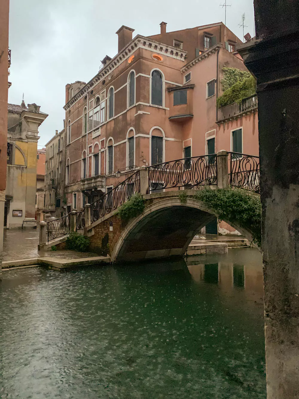 Venice canals at dusk with soft rain rippling across the water, reflecting the glow of lanterns and historic buildings, as gondolas glide silently through the moody, romantic atmosphere.