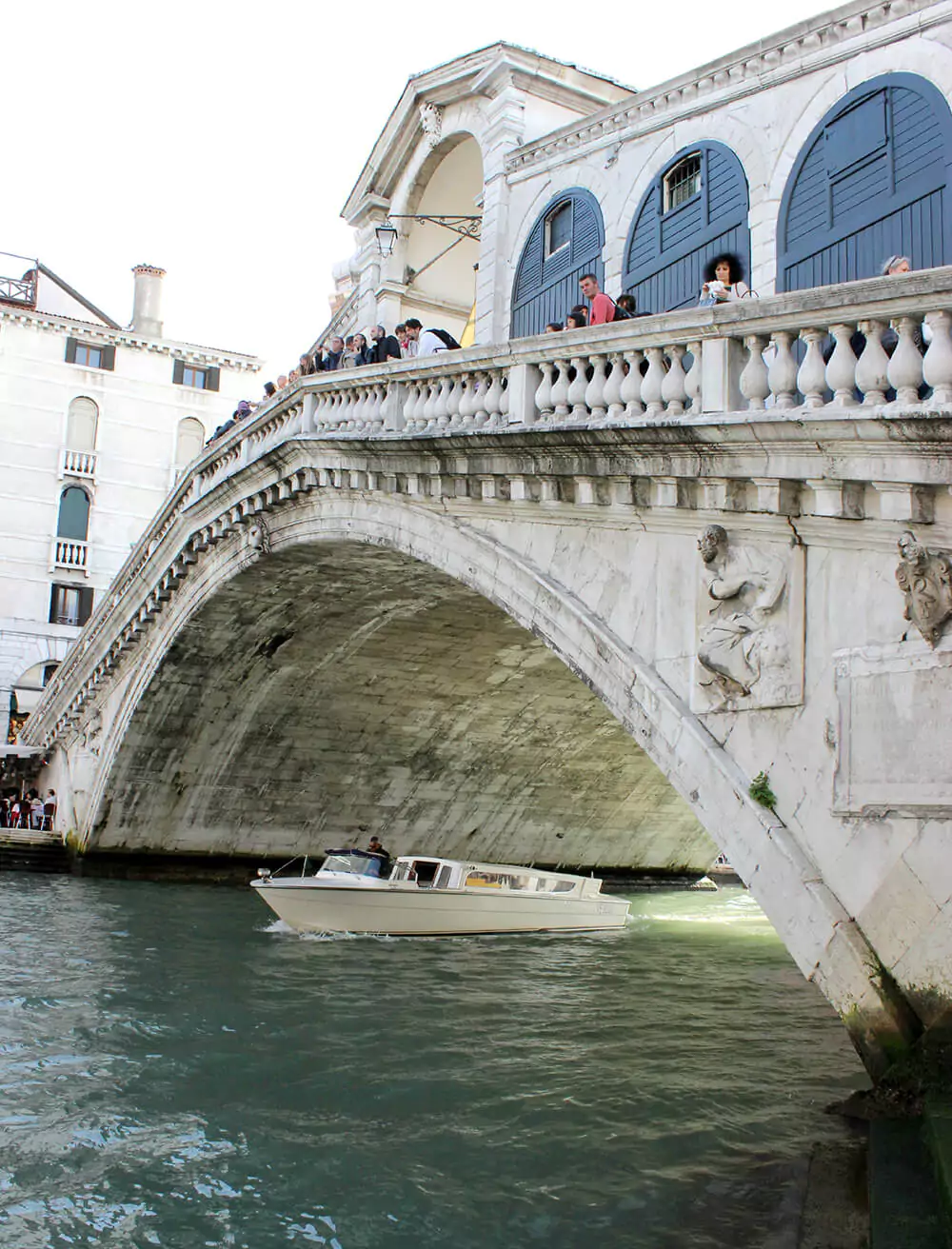 Tourist boat passing under the Rialto Bridge in Venice, Italy, with visitors taking photos and enjoying the scenic ride along the Grand Canal, framed by the bridge’s iconic stone arches.