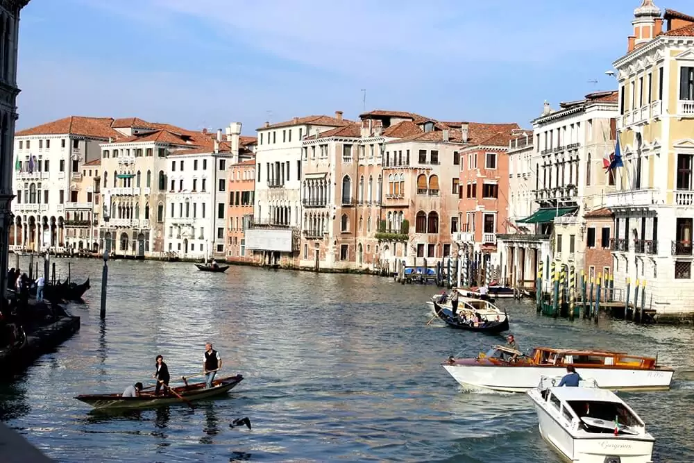 ondolas and boats gliding along the Grand Canal in Venice, Italy, surrounded by historic buildings with ornate facades. The lively waterway reflects the city’s charm and timeless atmosphere under a bright sky.