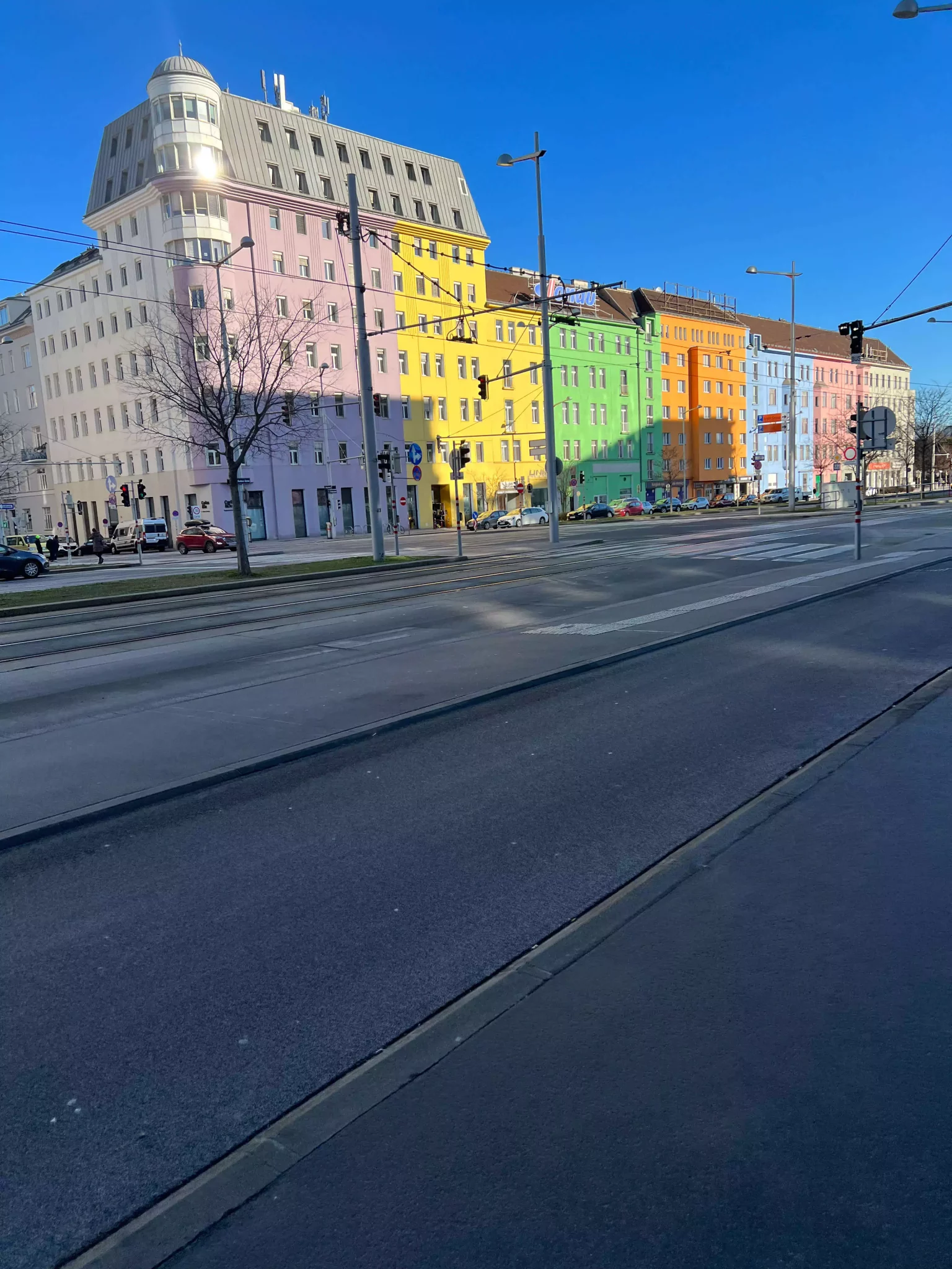 A row of colorful houses across from the train station in Vienna’s Quartier Belvedere district, each painted in soft pastel tones like peach, mint, and yellow. The cheerful facades add charm and contrast to the modern urban surroundings.