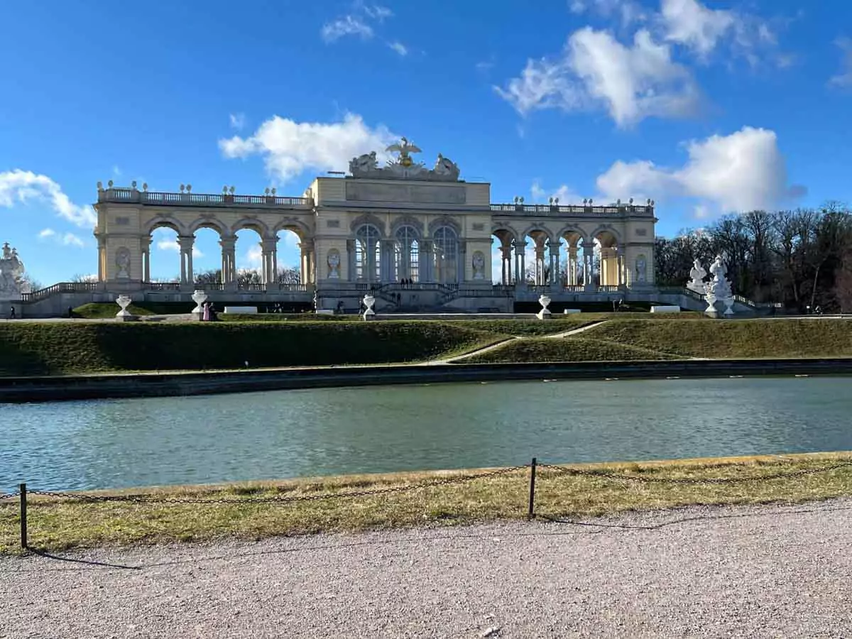 The Gloriette at Schönbrunn Palace in Vienna, Austria, standing atop a hill with its neoclassical columns reflected in the still pond below, surrounded by manicured lawns and garden paths.