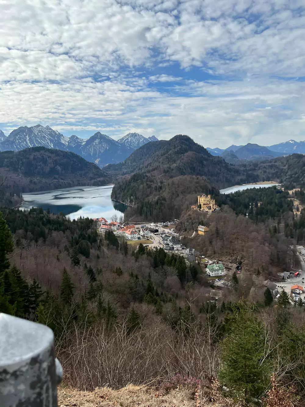 Hohenschwangau Castle perched on a wooded hill in Bavaria, Germany, overlooking a picturesque alpine valley with forested slopes, winding roads, and serene lakes in the distance.
