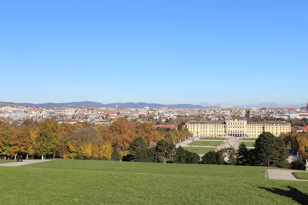 A panoramic view of Schönbrunn Palace in Vienna, Austria, with its grand yellow façade and manicured gardens stretching out below. In the background, the city of Vienna unfolds beneath a clear sky, blending imperial history with urban charm.