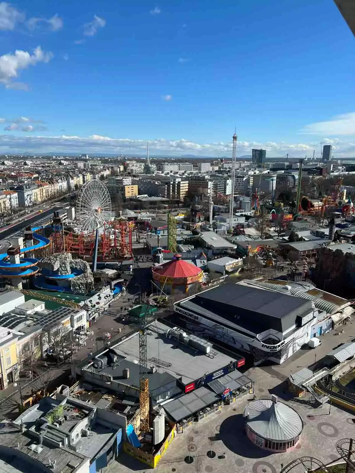 Panoramic view of Vienna’s Prater amusement park from the top of the Giant Ferris Wheel, featuring colorful rides, roller coasters, tree-lined paths, and the cityscape stretching into the distance.