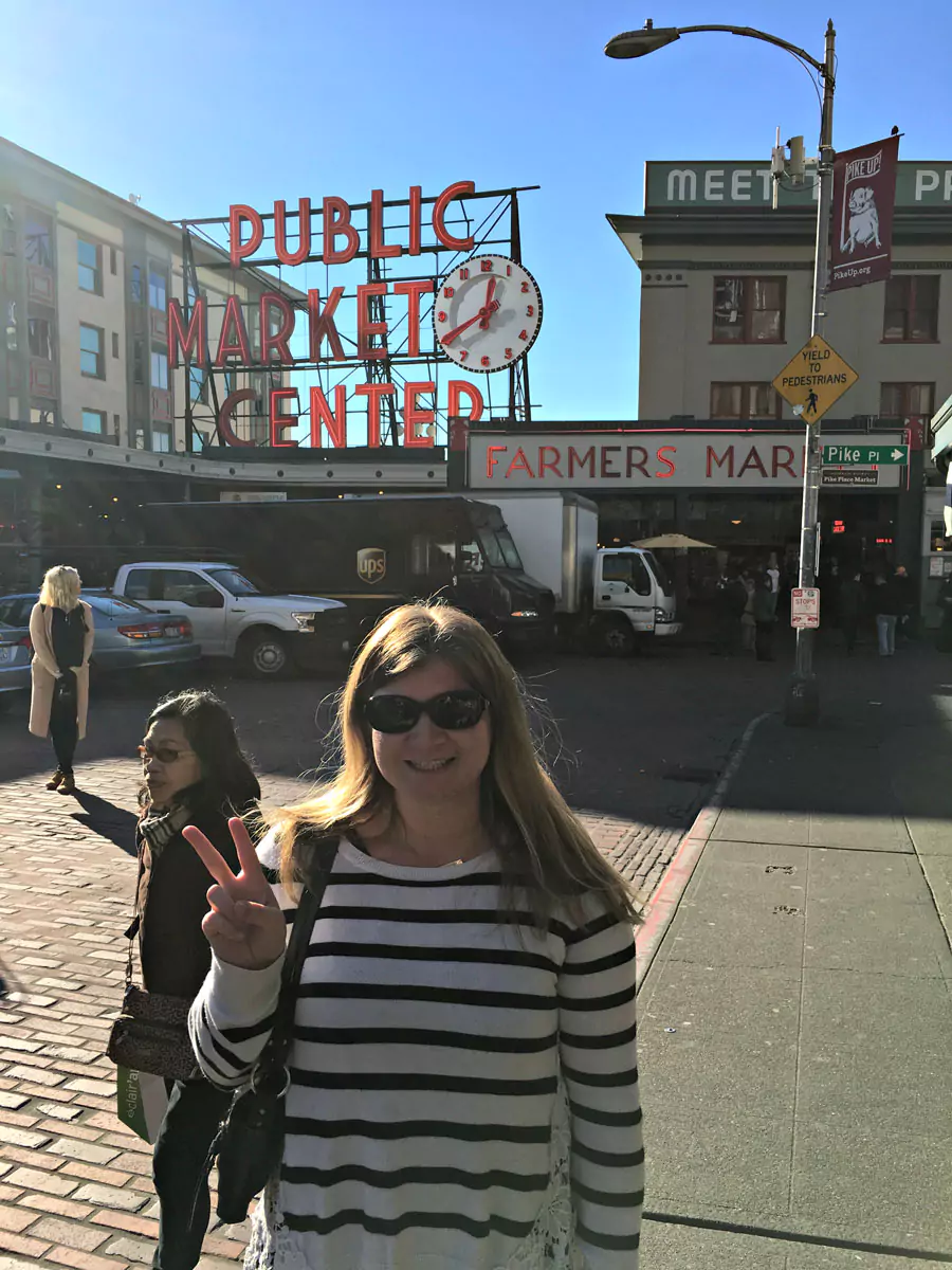 Katie at Pike Place Market in Seattle, smiling in front of the iconic red neon “Public Market Center” sign. The lively scene includes flower stands, local vendors, and the market’s classic urban energy.