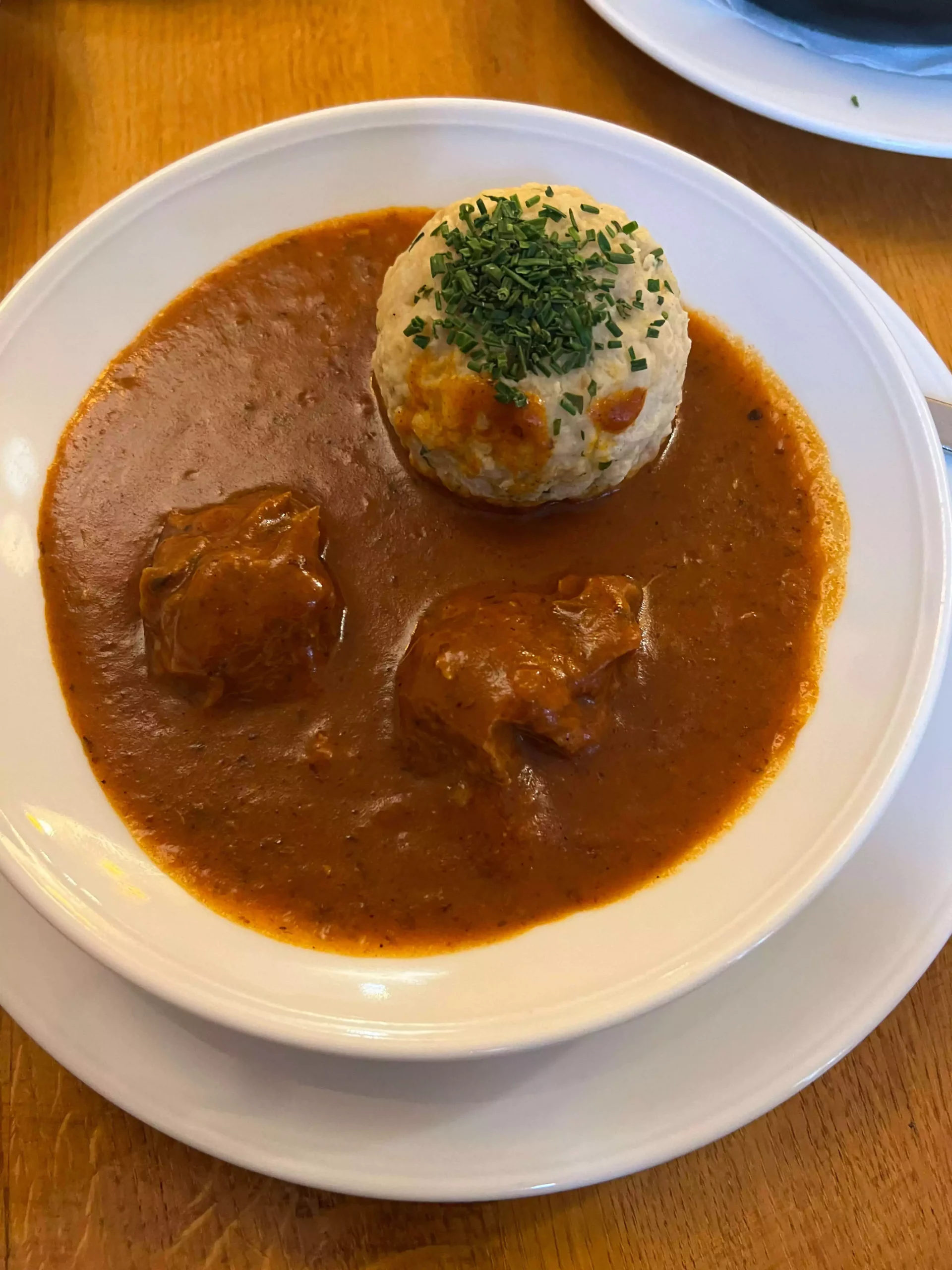 A white bowl filled with hearty goulash sits on a wooden table at Augustiner Bräugasthof Krimpelstätter in Salzburg, Austria. The rich, paprika-colored stew.