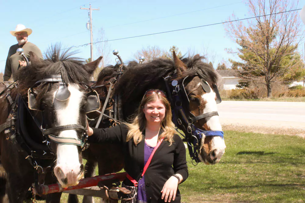 Katie stands next to a horse-drawn carriage outside Lautenbach's Orchard Country Winery & Market in Door County, Wisconsin. The rustic setting includes orchard trees and charming market buildings, capturing a quaint countryside experience.
